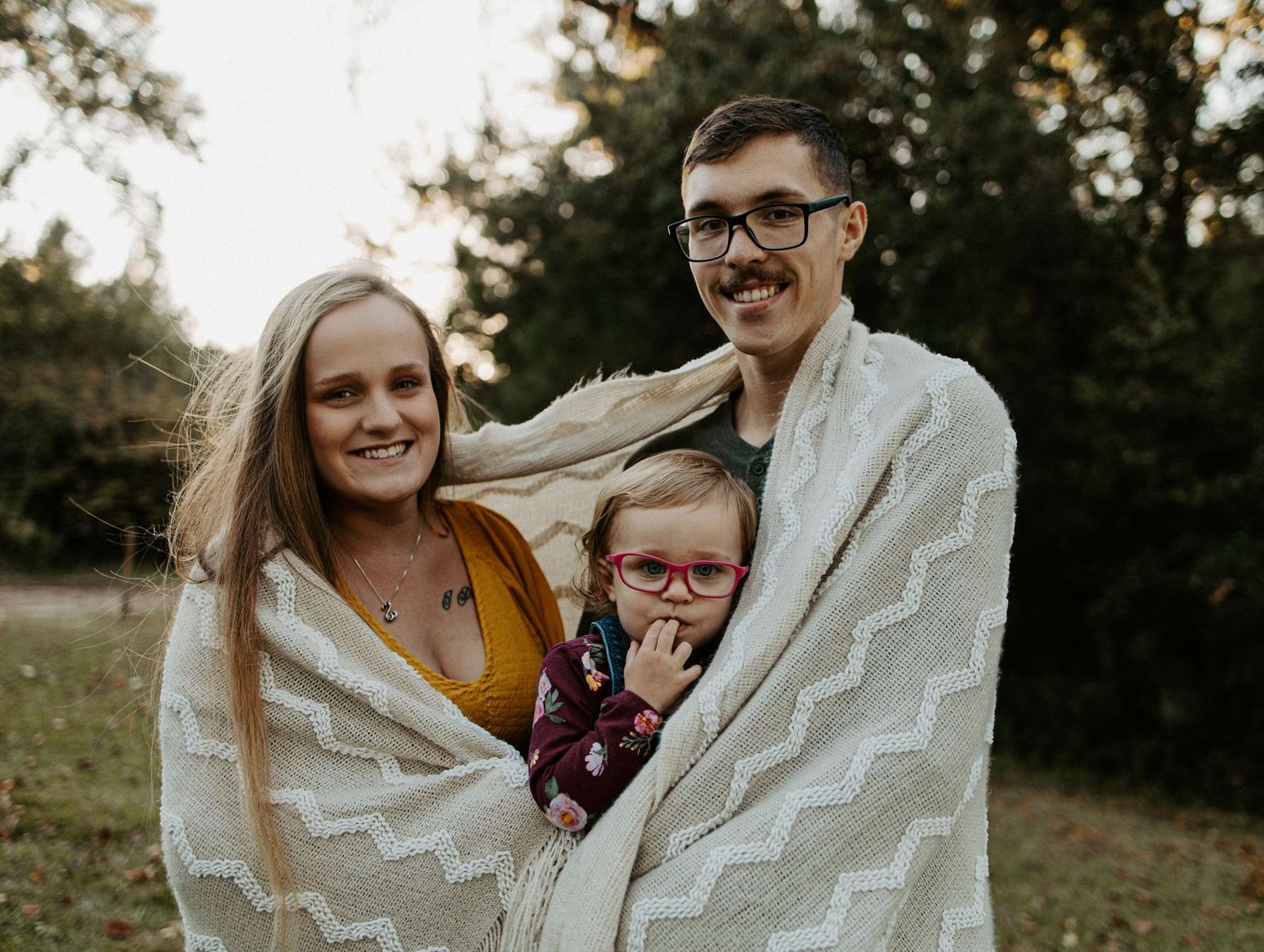 Smiling family wrapped in a cozy white blanket with their toddler in glasses outdoors at golden hour.