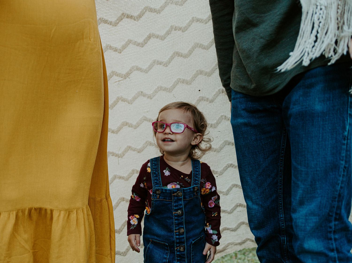 Toddler girl in overalls and pink glasses smiling up between two adults during a family photo session.