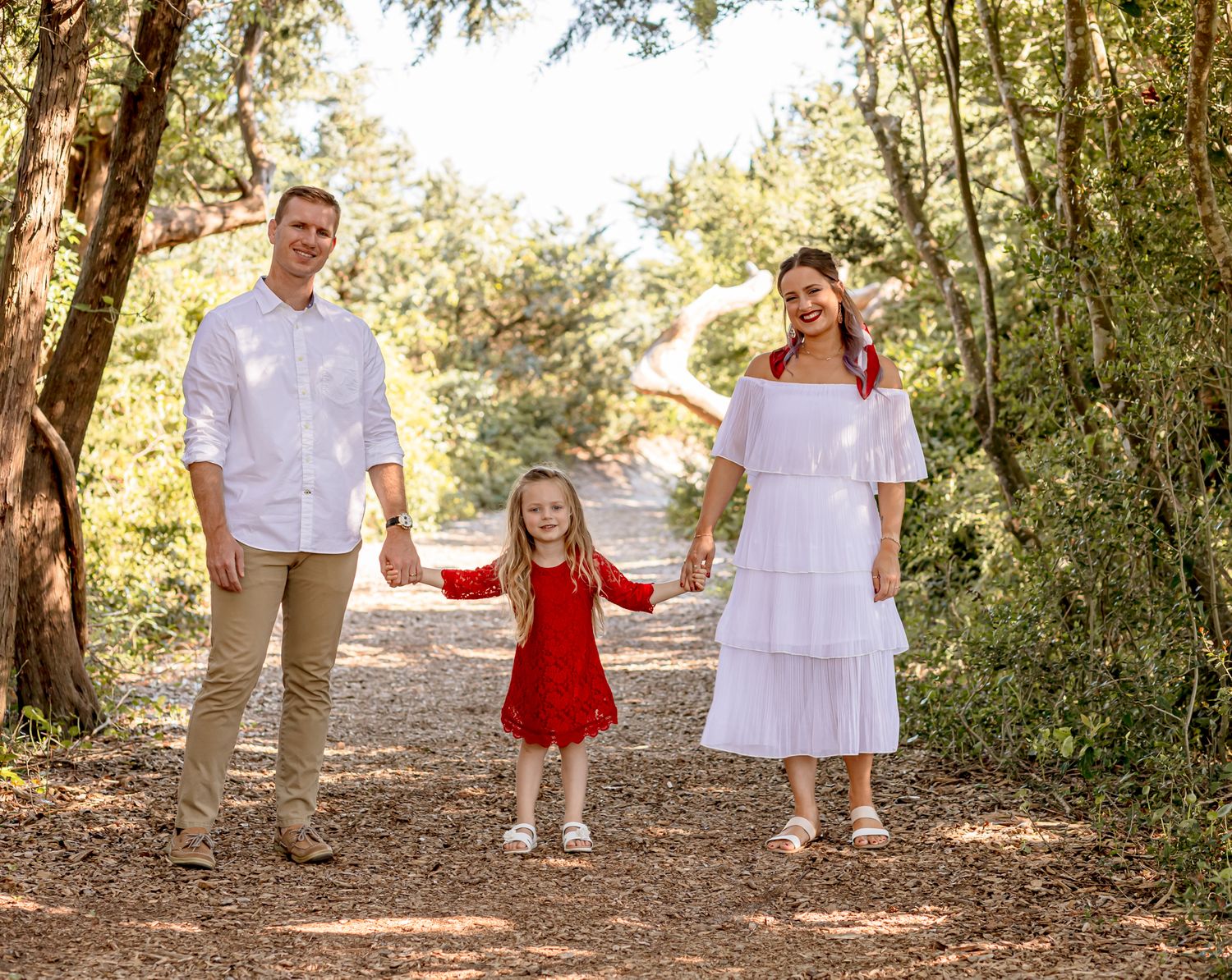 Happy family of three holds hands on a sunny tree-lined path, parents in white, child in red.