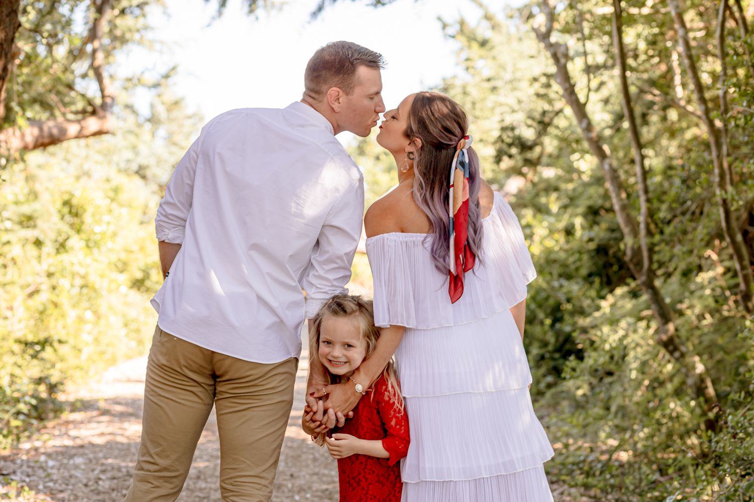 Parents share a kiss while their young daughter in red dress giggles between them outdoors.