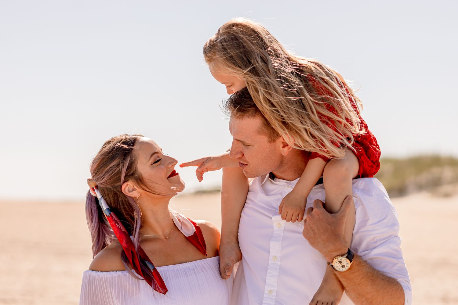 Cheerful family moment on beach as father carries daughter on shoulders while mother smiles.