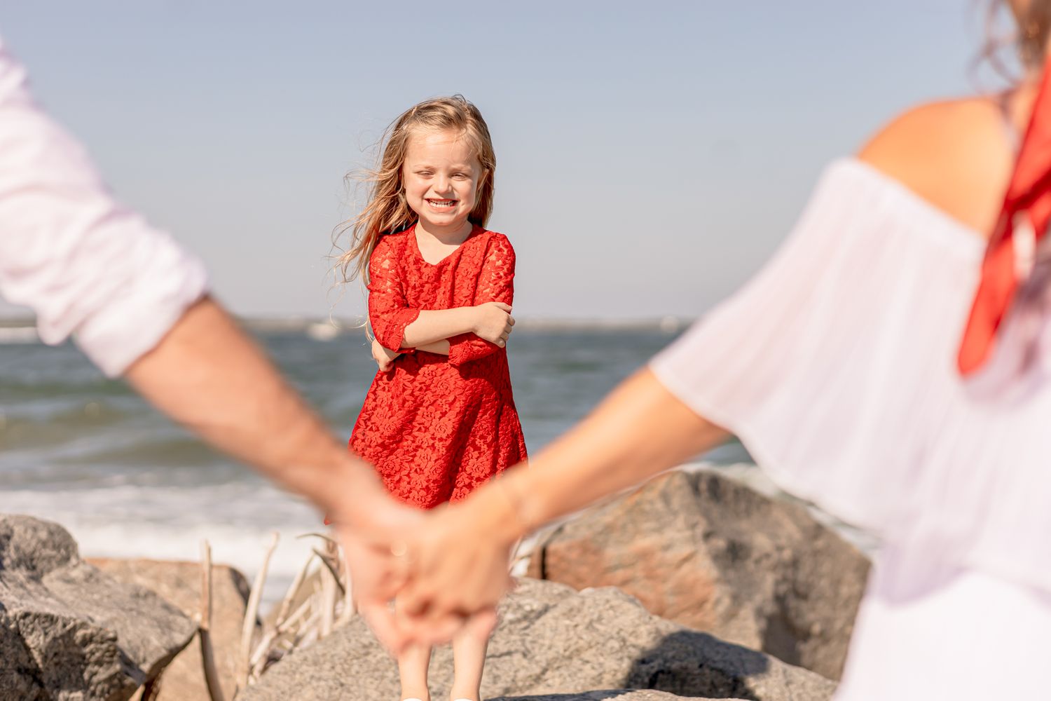 Smiling little girl in red dress stands on rocks as parents hold hands in the foreground.