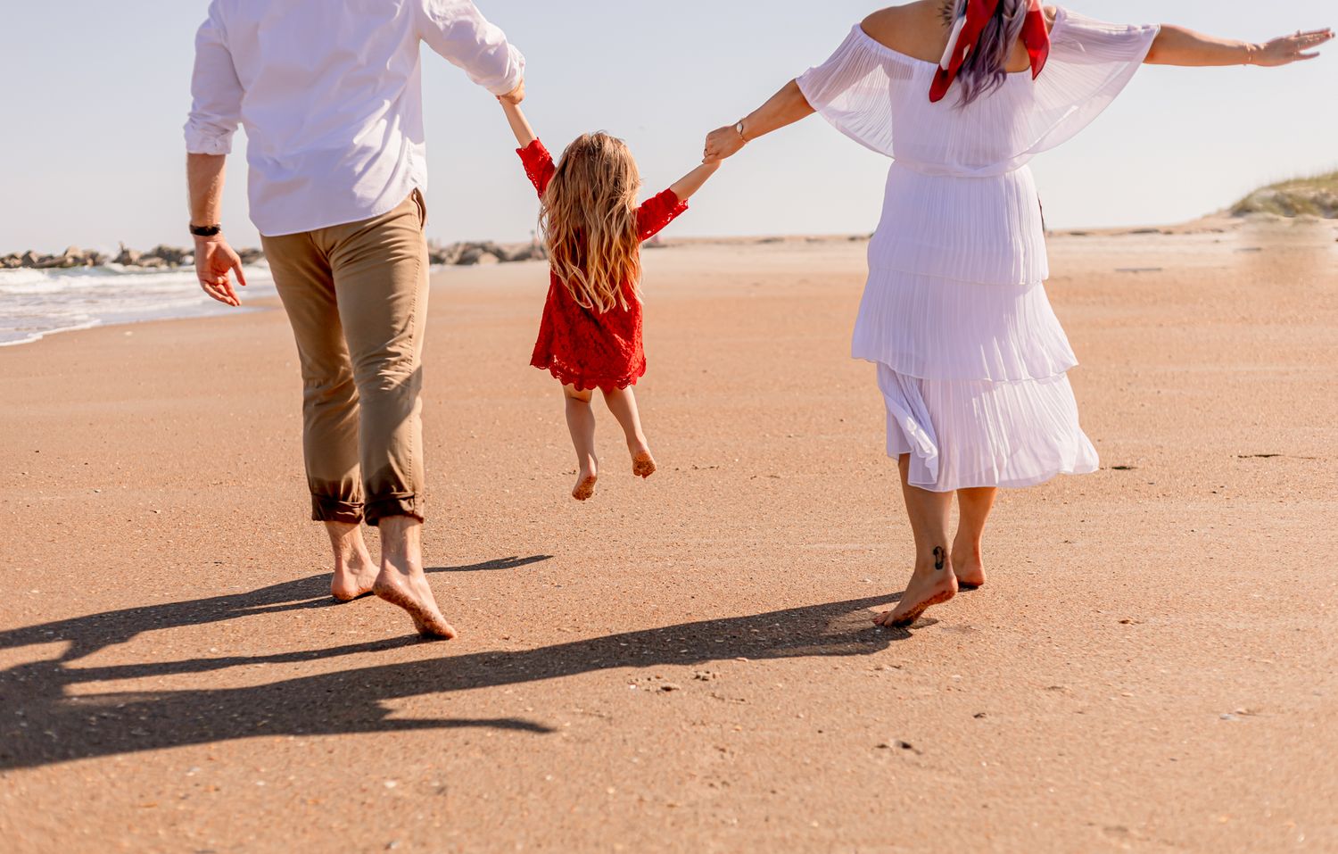 Family of three walks barefoot on sandy beach, swinging young daughter in red dress between them.