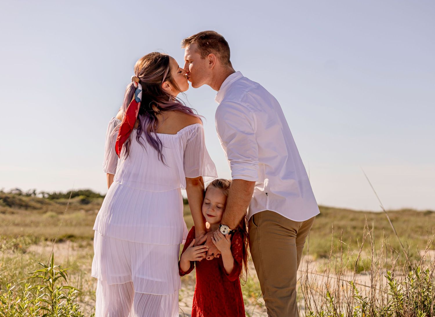 Couple kisses while their young daughter in red dress hugs them in a sunlit grassy field.