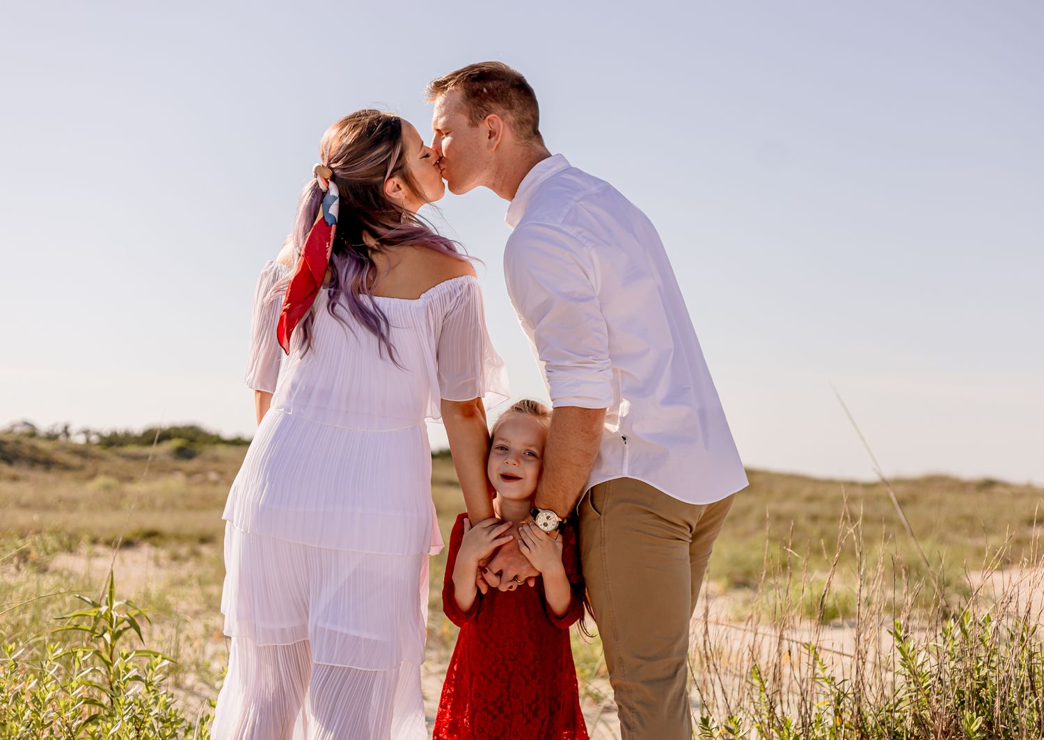 Parents share a kiss while young daughter in red dress stands between them in sunny meadow.