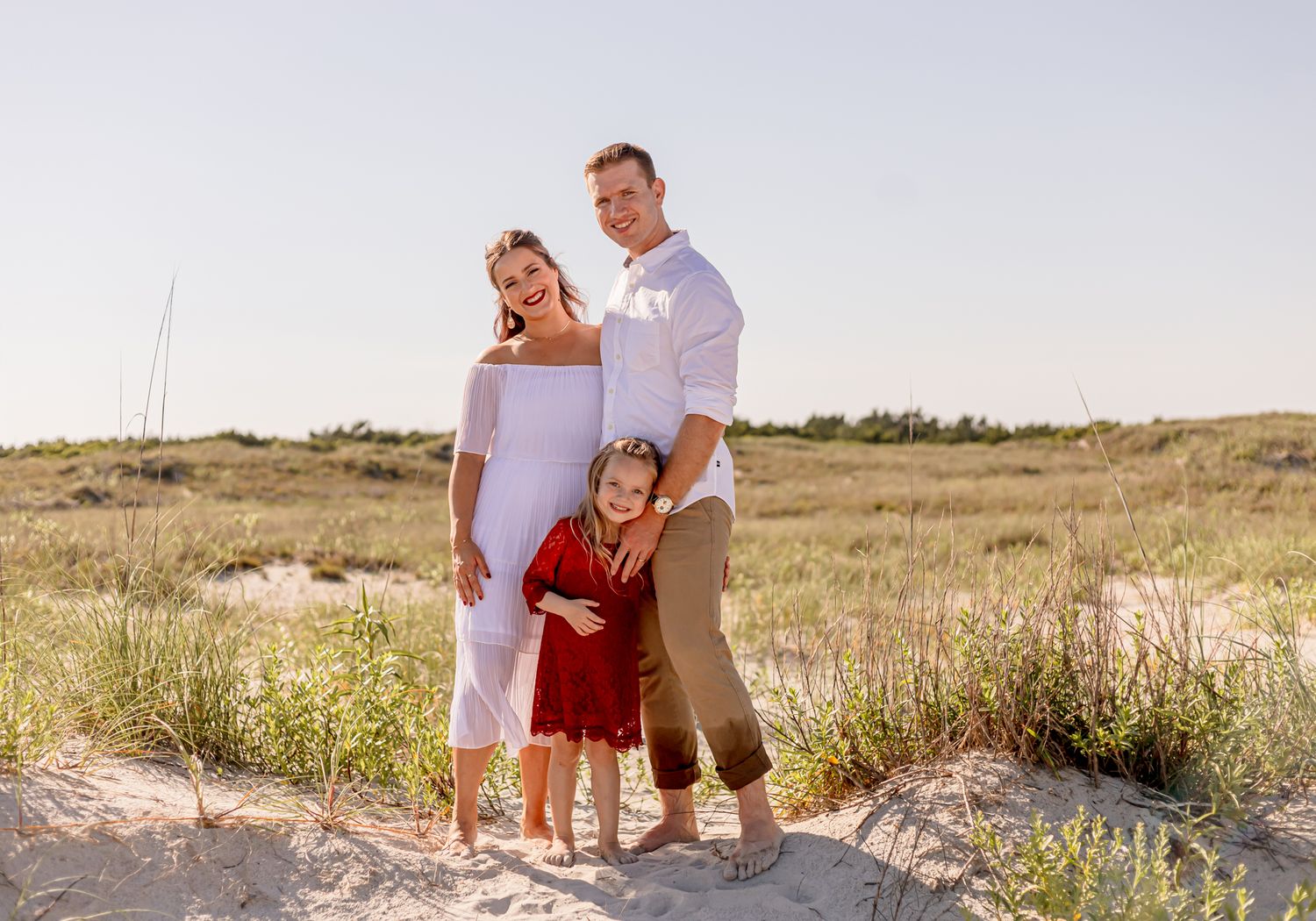Family of three posing on sandy beach dunes, pregnant mother in white dress with daughter in red dress.