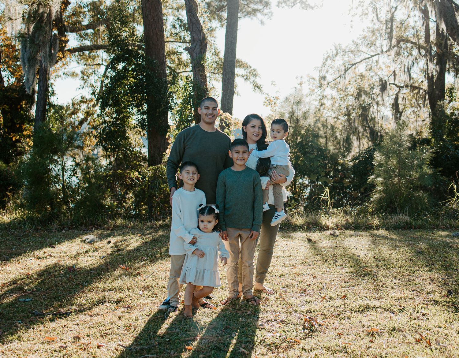 Family of five standing together in a sunlit wooded area with Spanish moss trees during an outdoor portrait.