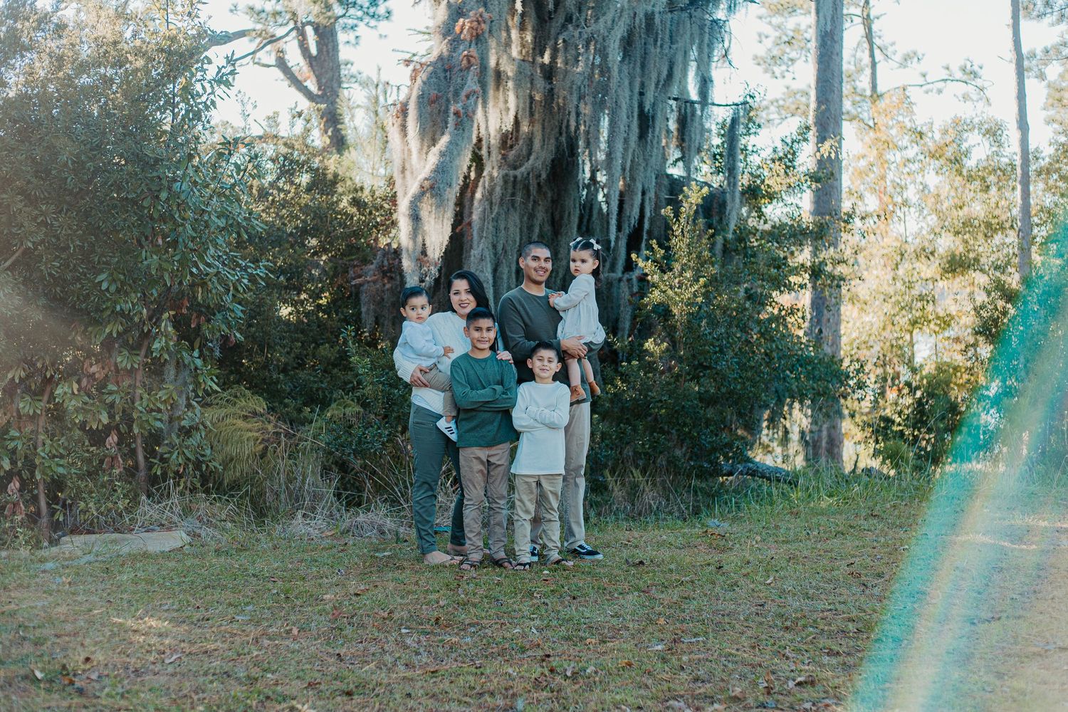 Family of six posing outdoors under a large moss-draped tree in a lush green setting.
