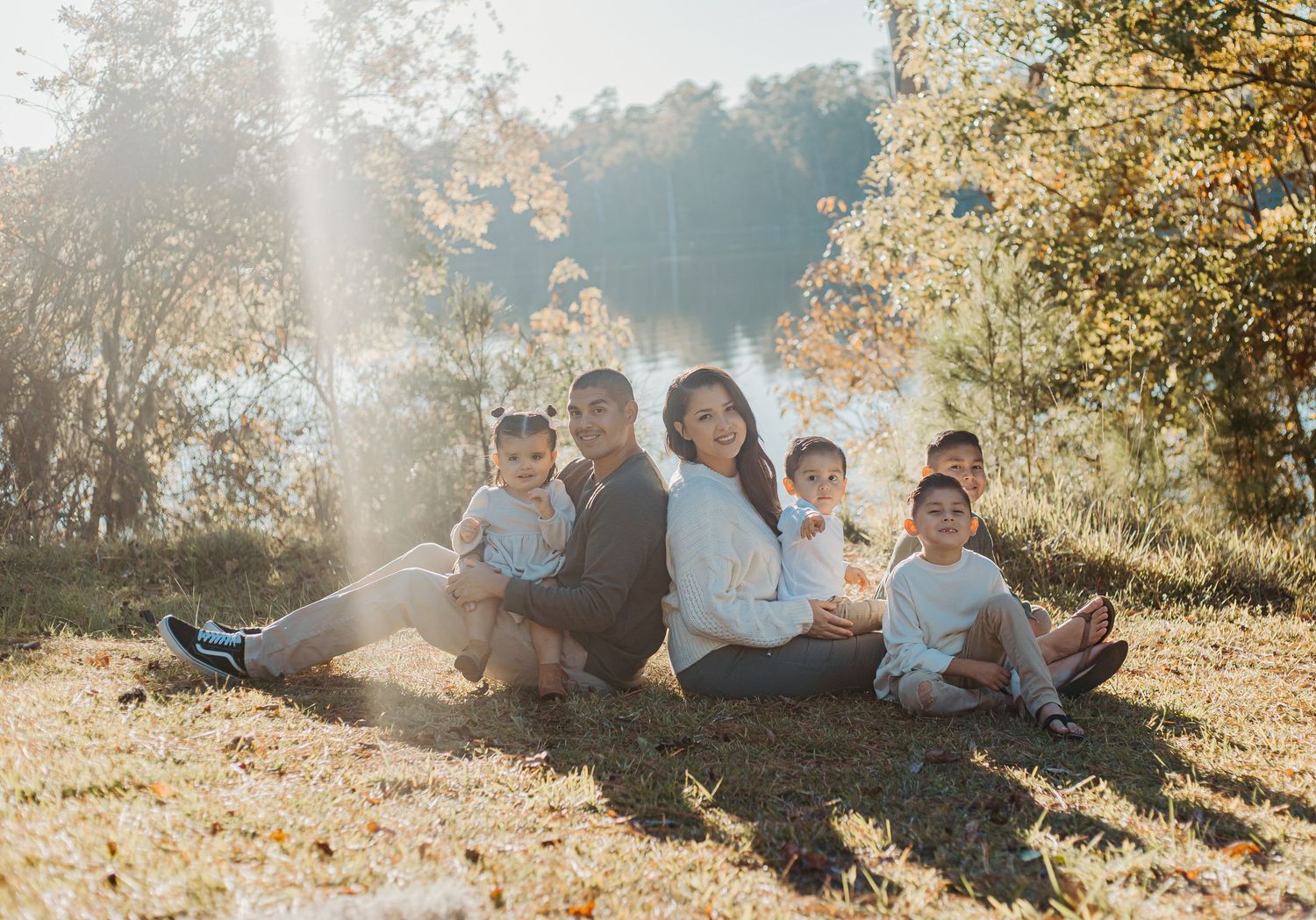 Family of six sitting on grass by a sunlit lake surrounded by autumn foliage.