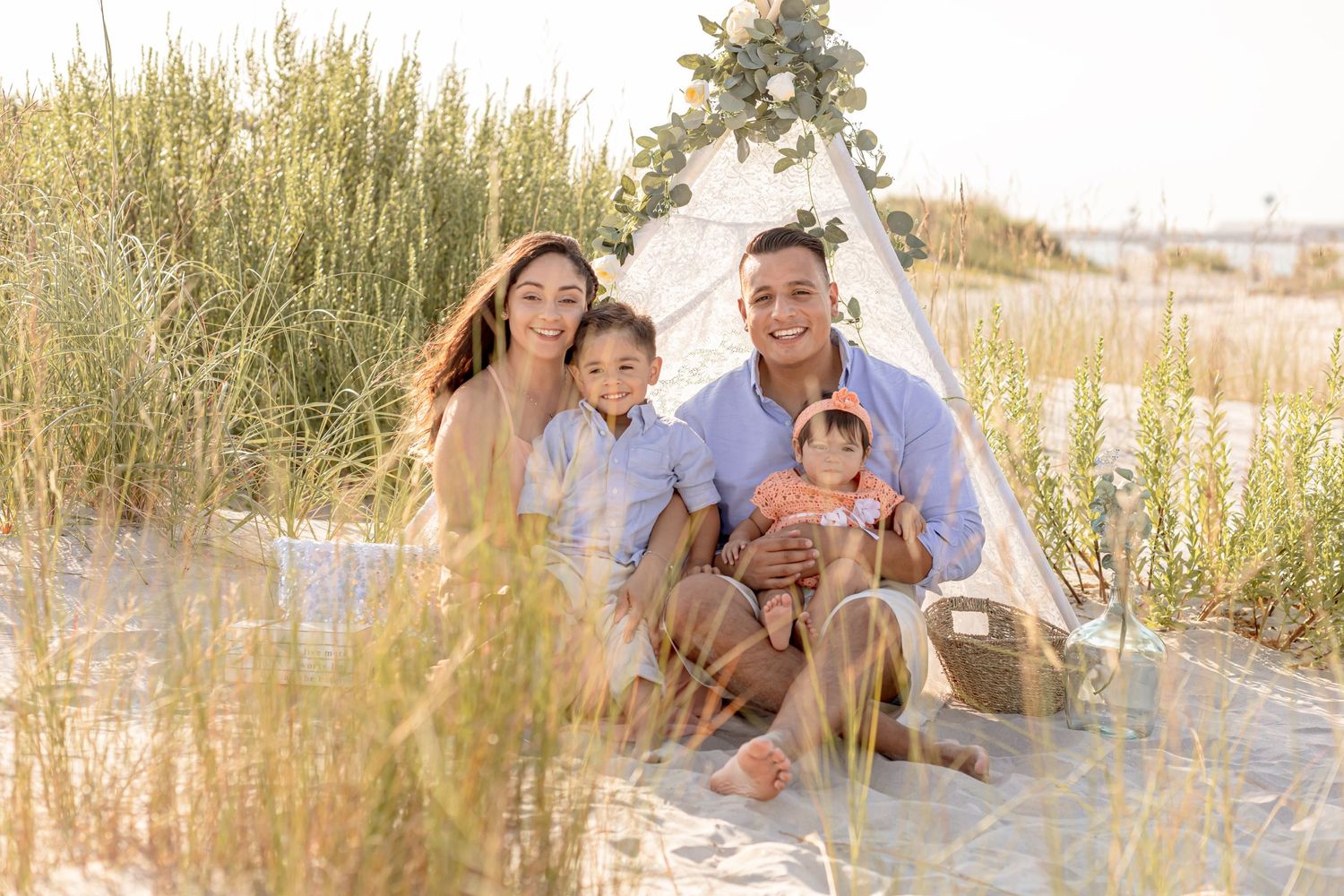 Happy family of four posing by a decorated teepee on sandy beach dunes at sunset.