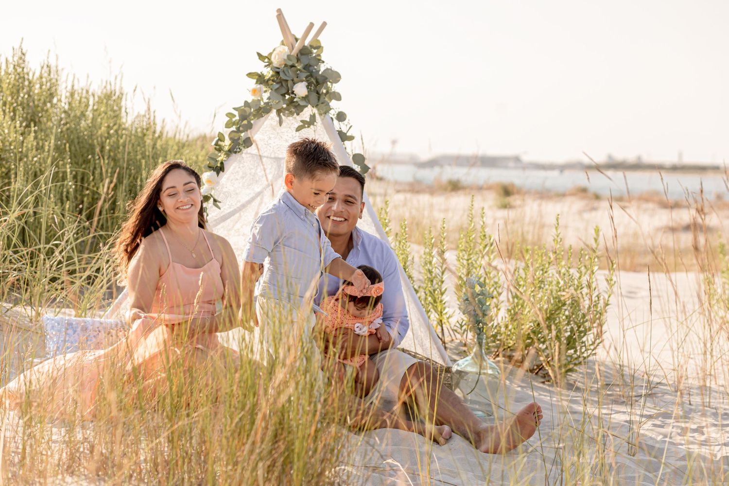 Family sitting on beach sand dunes near a boho teepee during golden hour photo session.