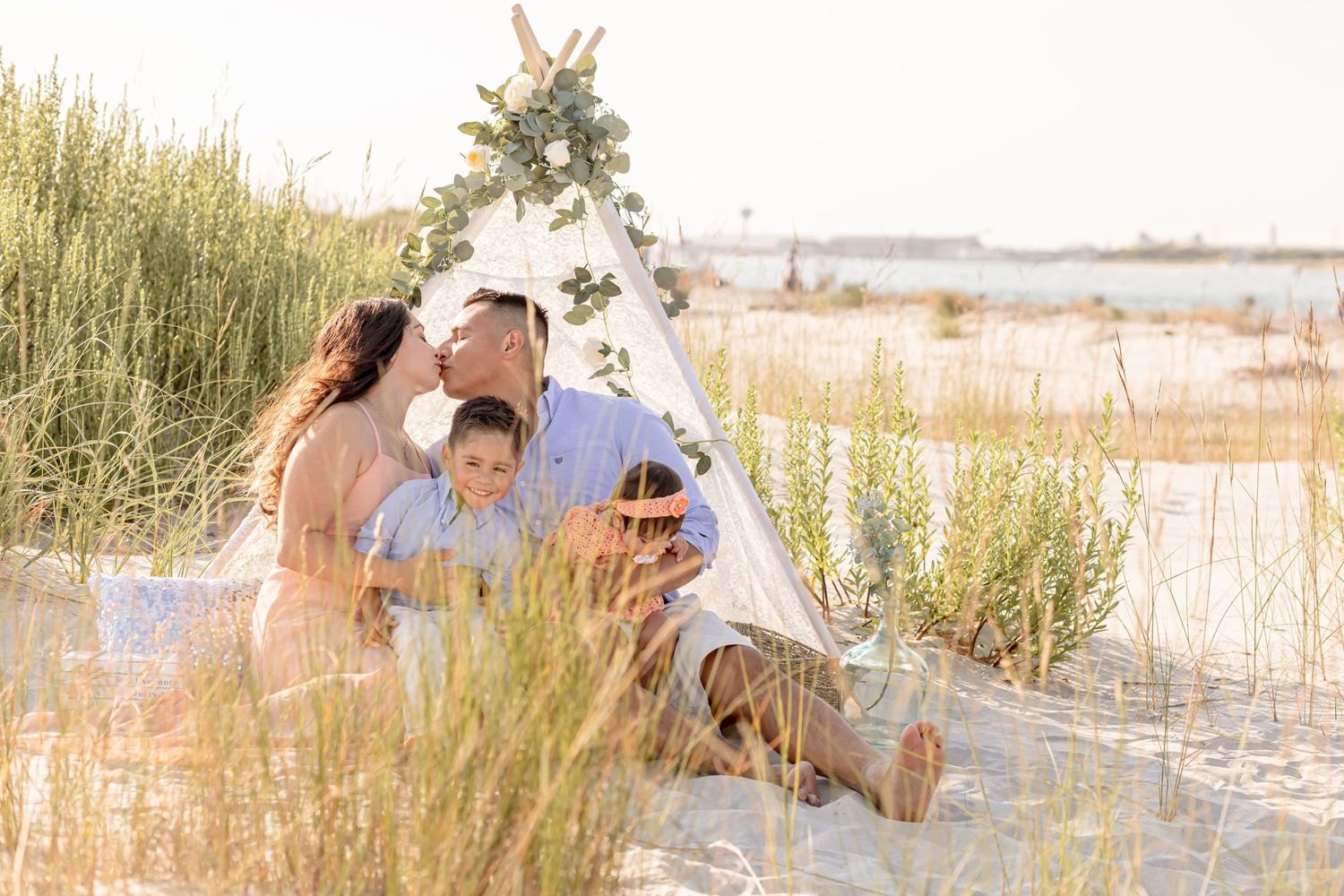 Family sharing tender moment together by a boho teepee on sandy beach dunes at sunset.