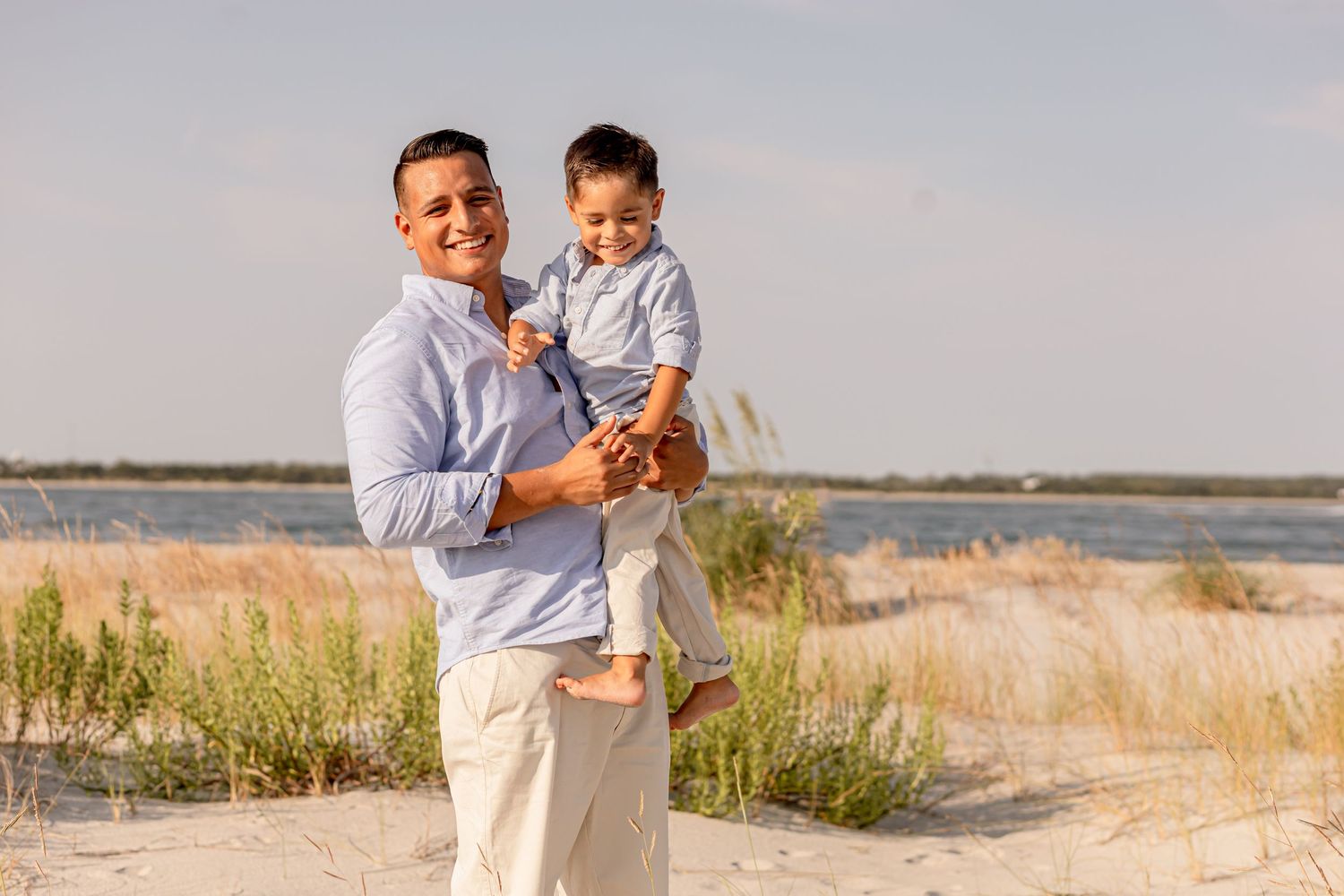 Smiling father holding young toddler son on a sunny beach with tall grass background.