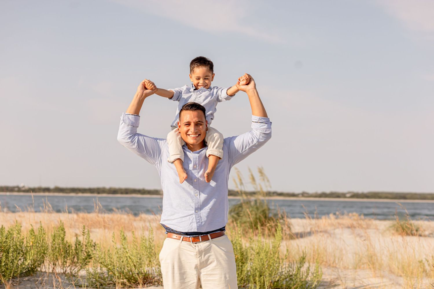 Joyful father carrying young son on shoulders on a sunny beach with water in background.