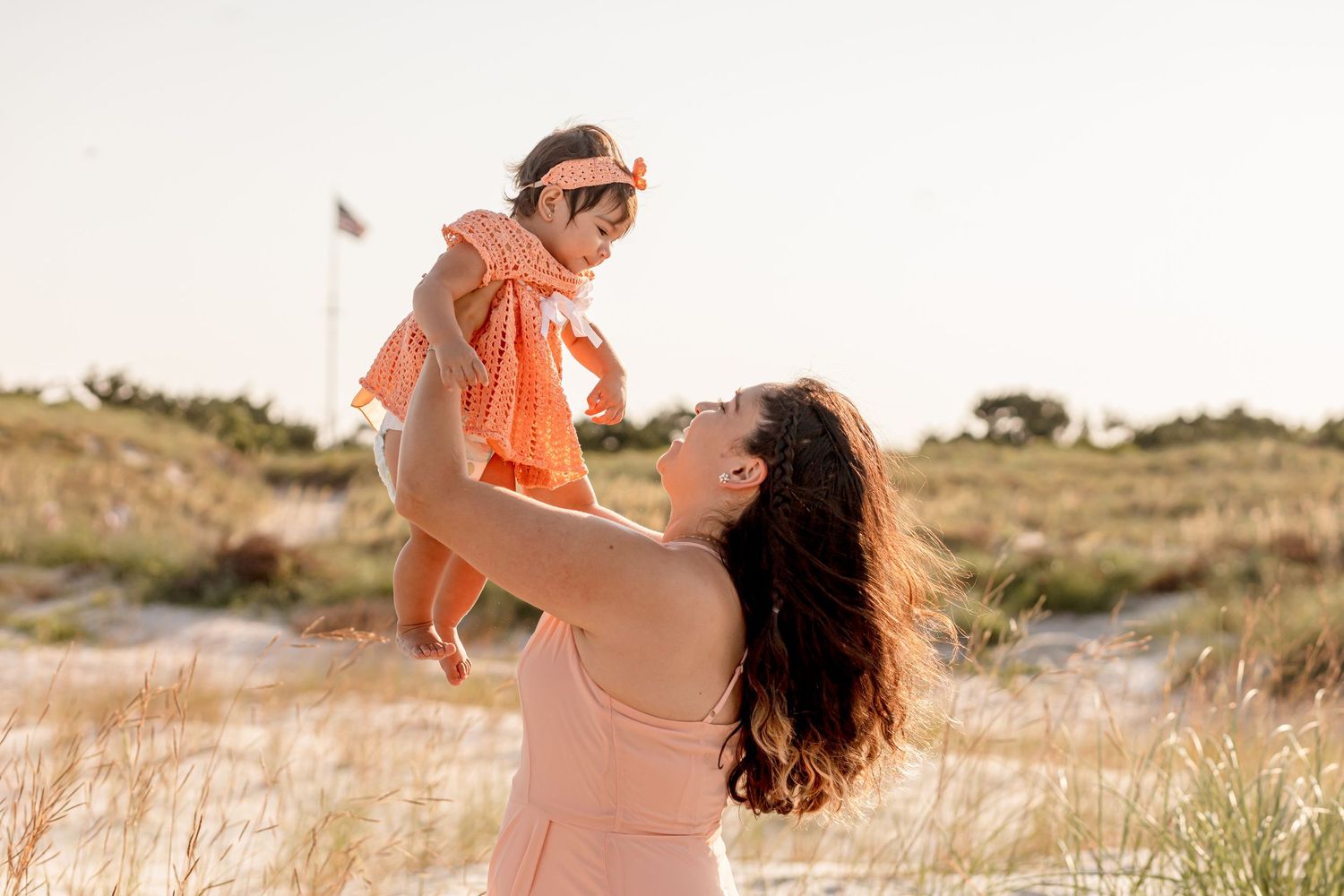 Loving mother lifting laughing baby girl in orange dress on a bright sandy beach.