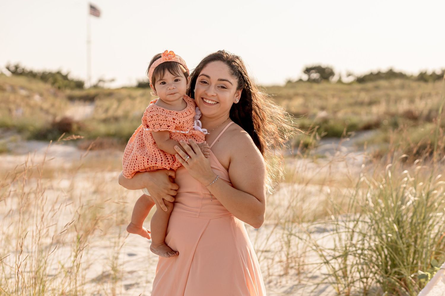 Smiling mother holding baby girl in matching orange outfits on a sunny beach setting.