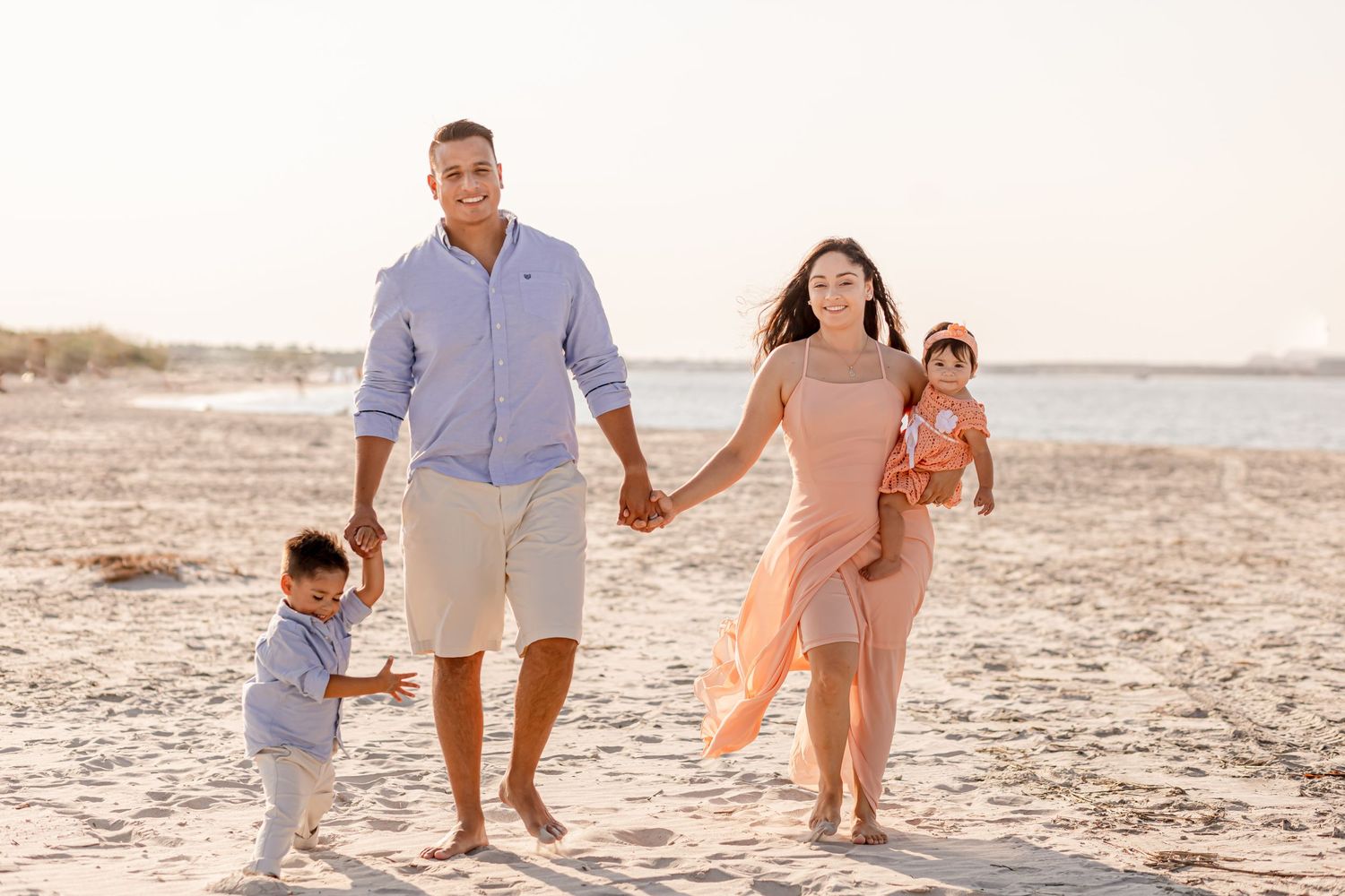 Smiling family of four strolling along a sunlit sandy beach holding hands together.