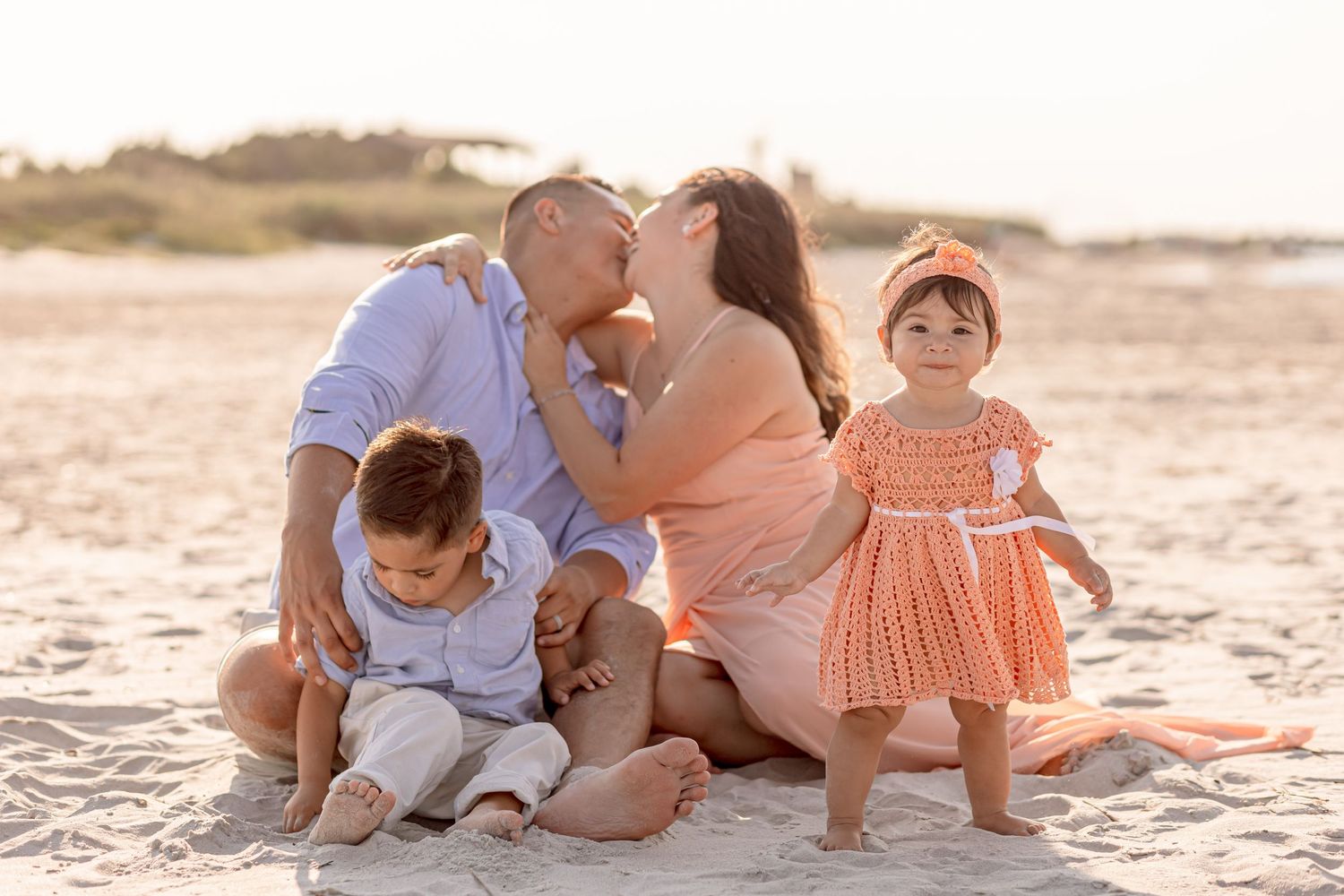 Family of four sitting on beach at golden hour, parents kissing while children play in sand.