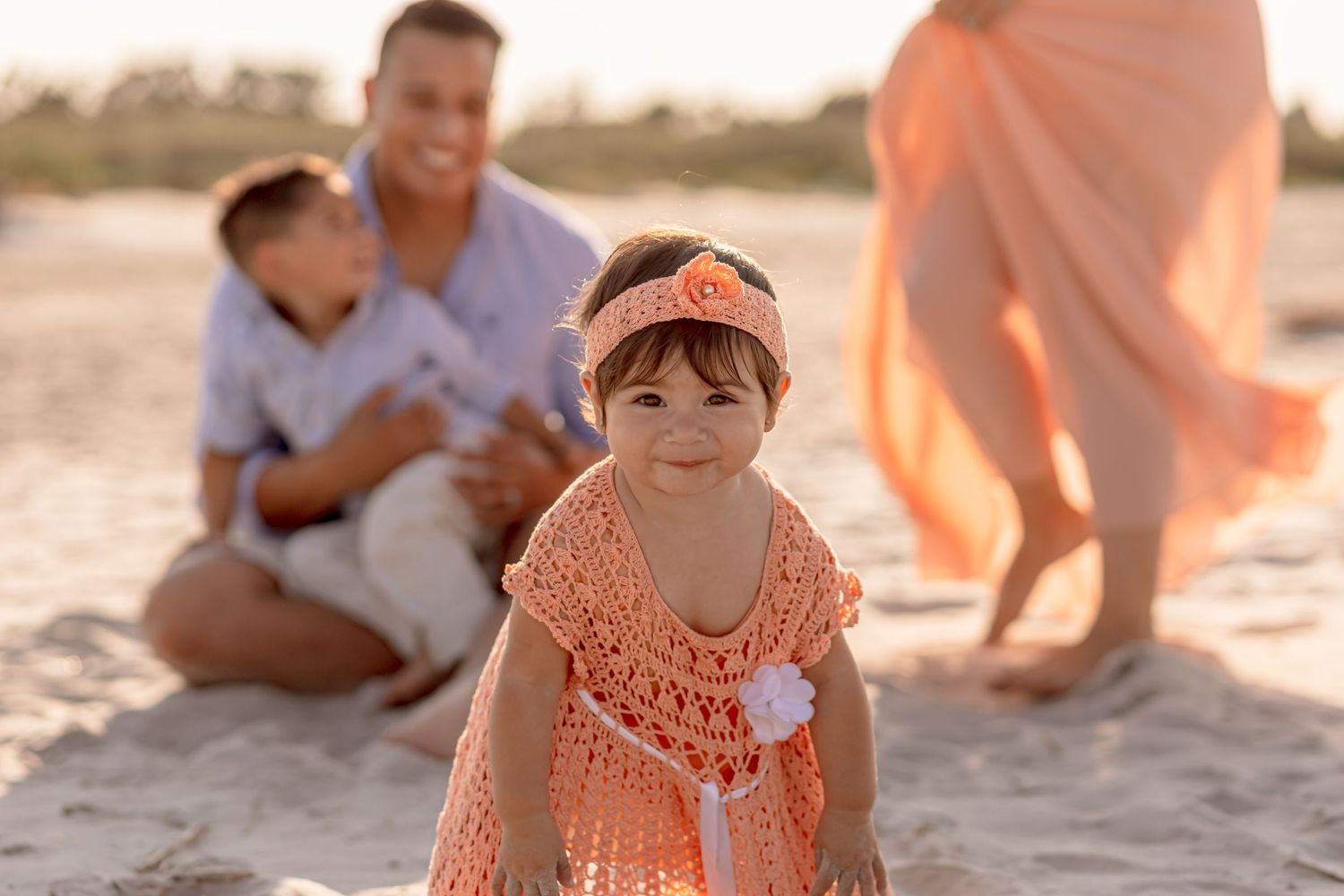 Adorable toddler girl in orange crochet dress with headband smiling on beach at sunset.