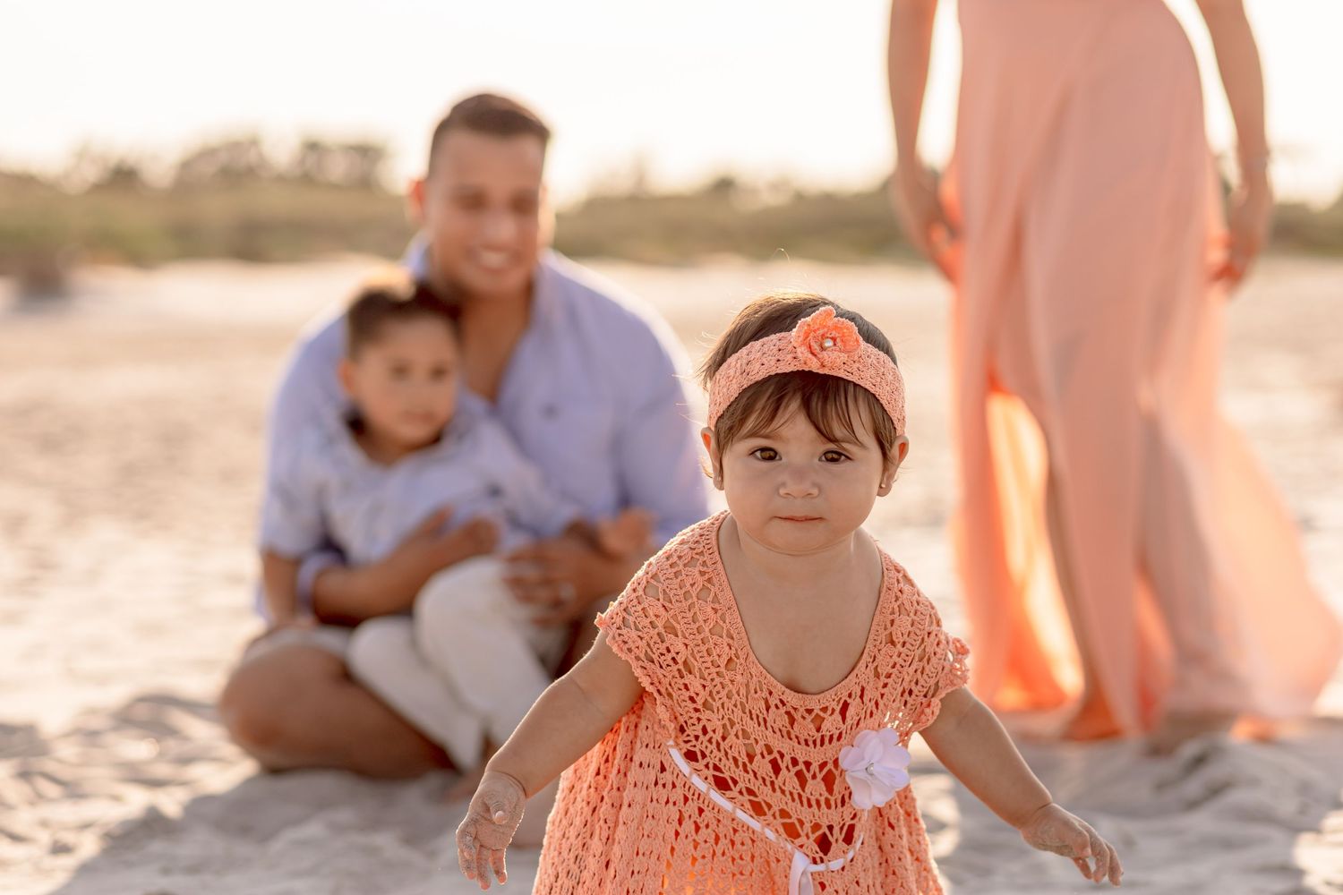 Cute baby girl in peach crochet dress crawling on beach with family sitting behind her.