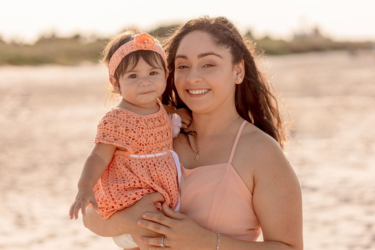 Smiling mother holding baby girl in matching peach outfits on sunny beach portrait.