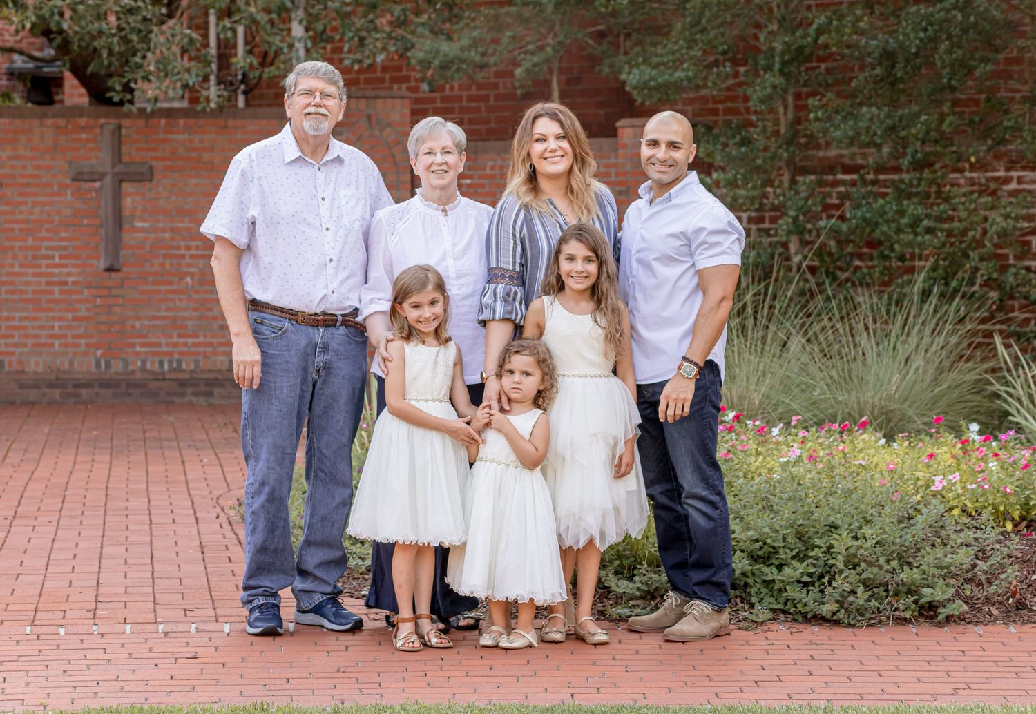 Extended family group photo with grandparents, parents, and three girls in white dresses by a brick church.