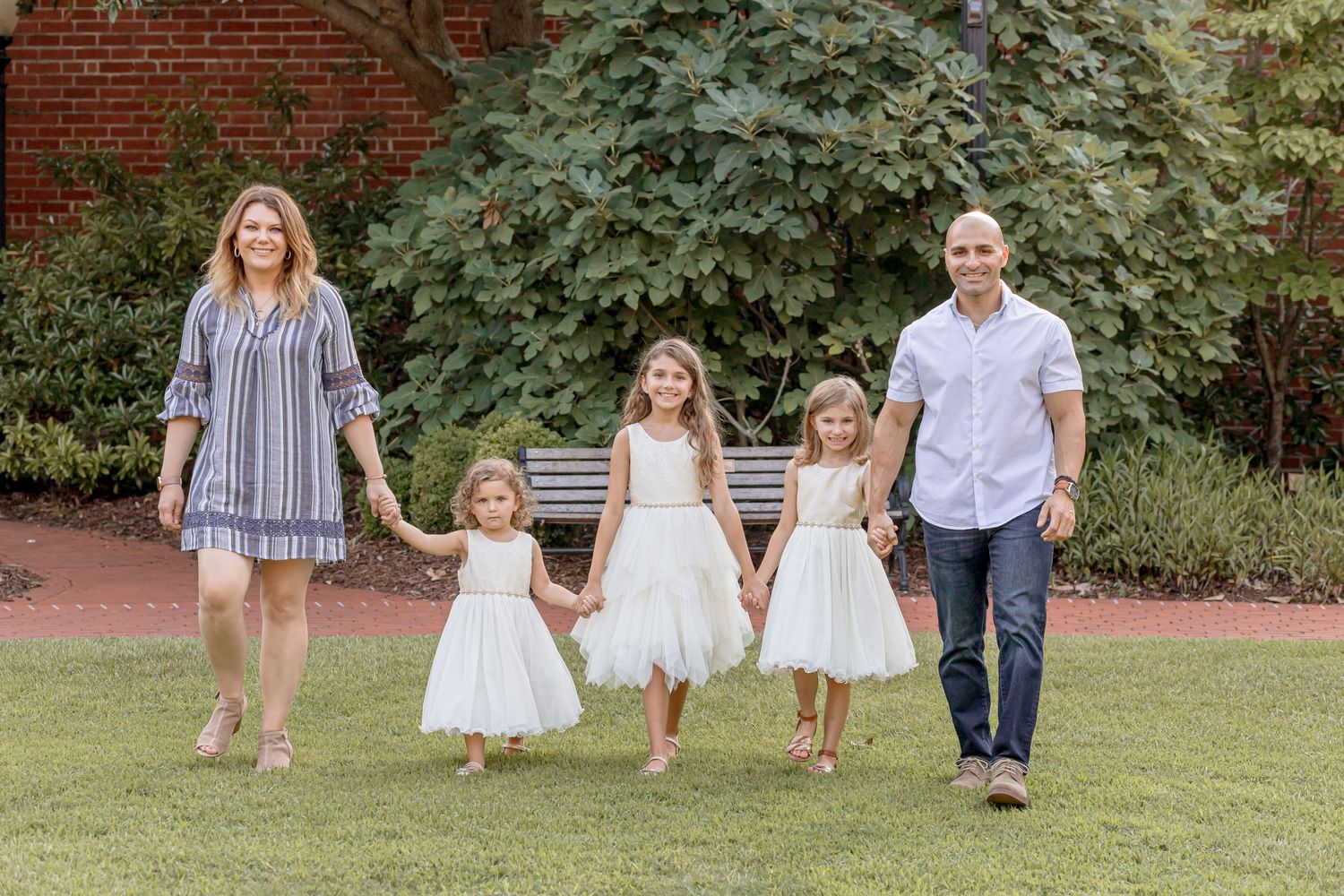 Family of five holding hands walking on green lawn, three girls in white dresses between their parents.