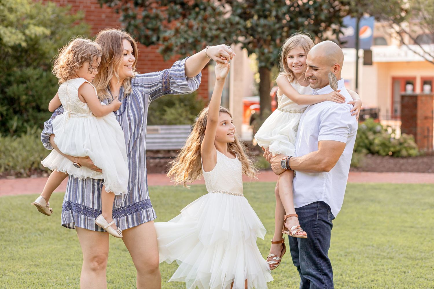 Joyful family of five playing outdoors, parents spinning and holding three daughters in white dresses.