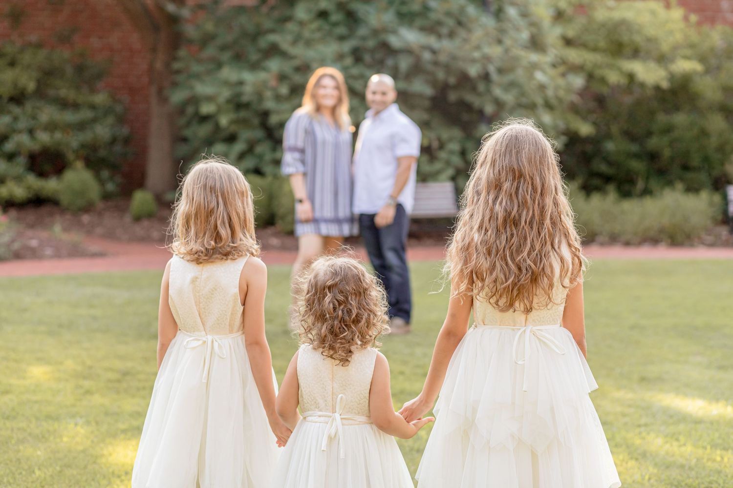 Three girls in white dresses holding hands, viewed from behind, facing their parents on a green lawn.
