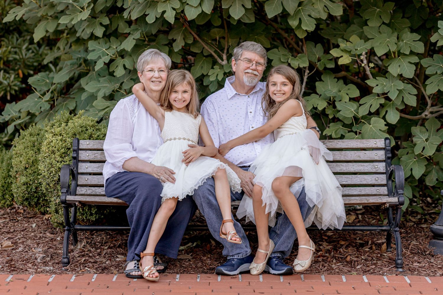 Grandparents sitting on a bench with two granddaughters in white dresses posing for a family portrait.