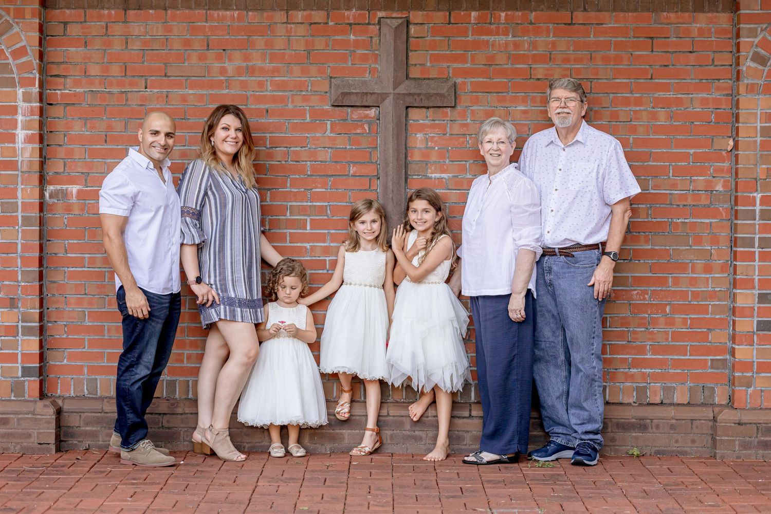 Extended family posing in front of a brick wall with a wooden cross, including three girls in white dresses.