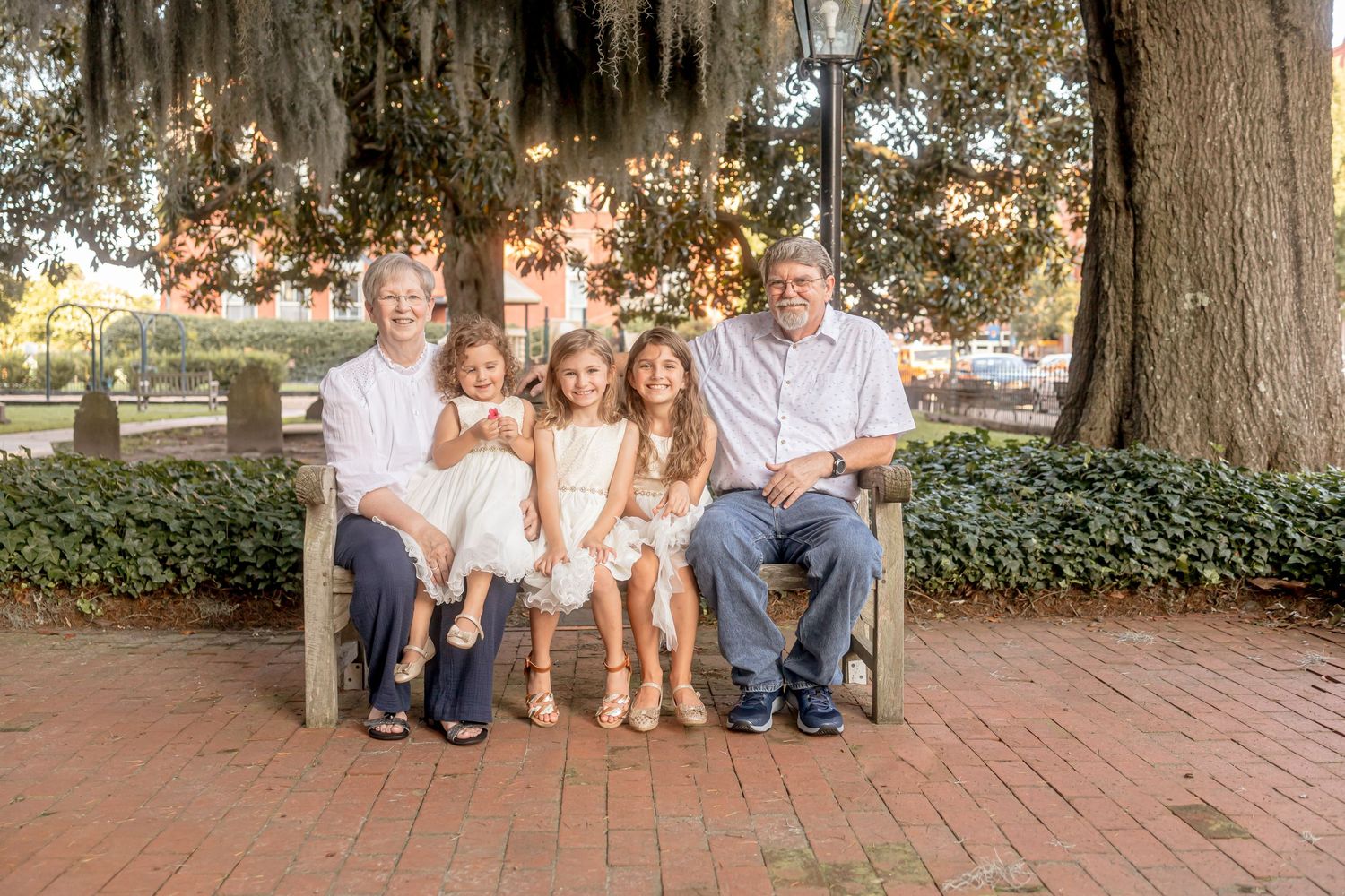 Grandparents sitting on a bench with three young girls in white dresses in a charming outdoor setting.