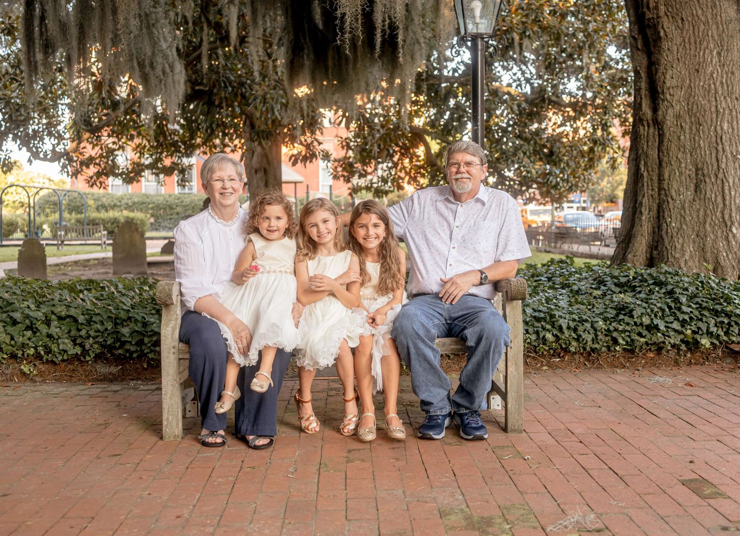Grandparents and grandchildren sitting on a bench under large trees on a brick plaza smiling together.