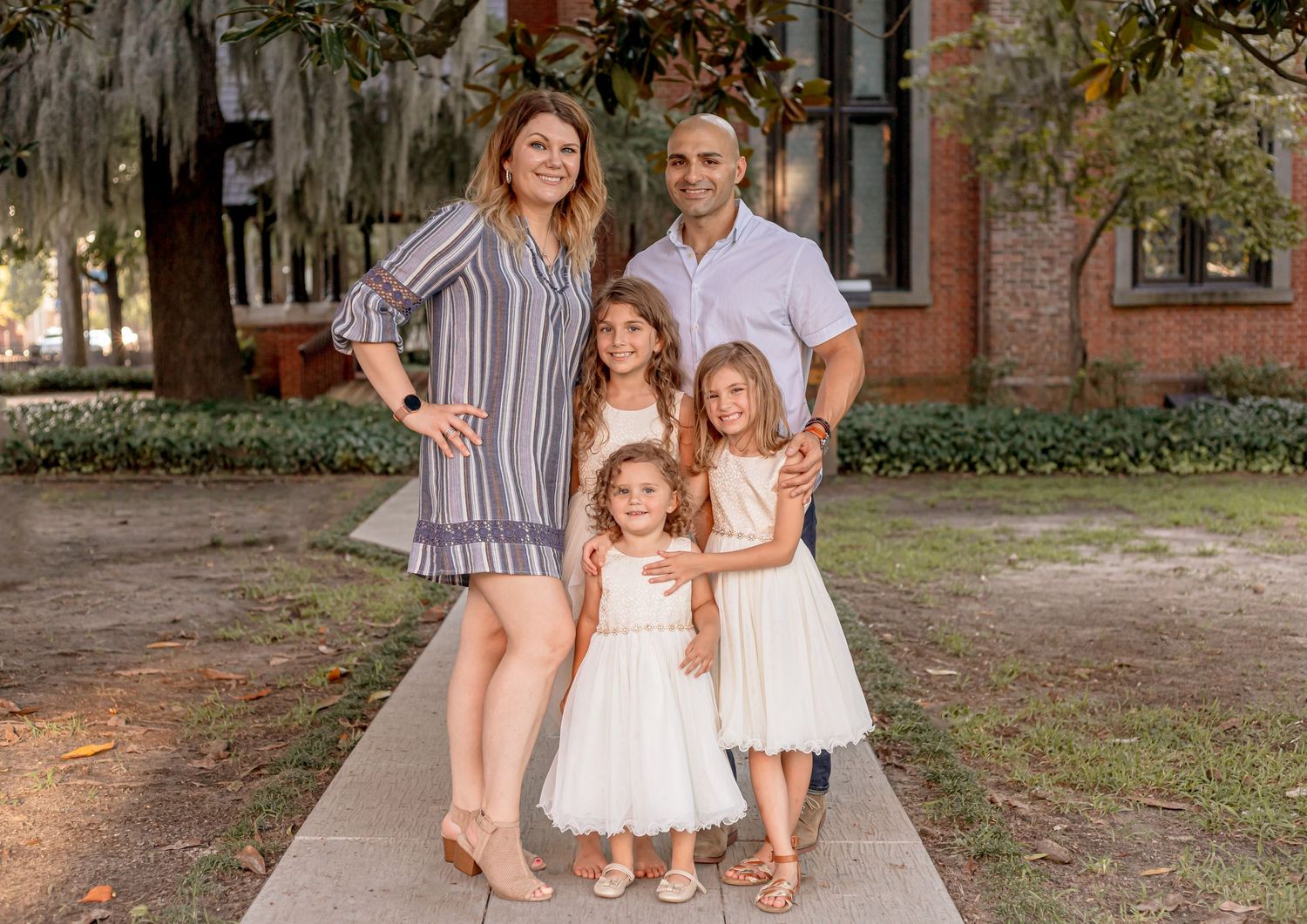 Family of five with three young girls in white dresses posing outdoors near a brick building.