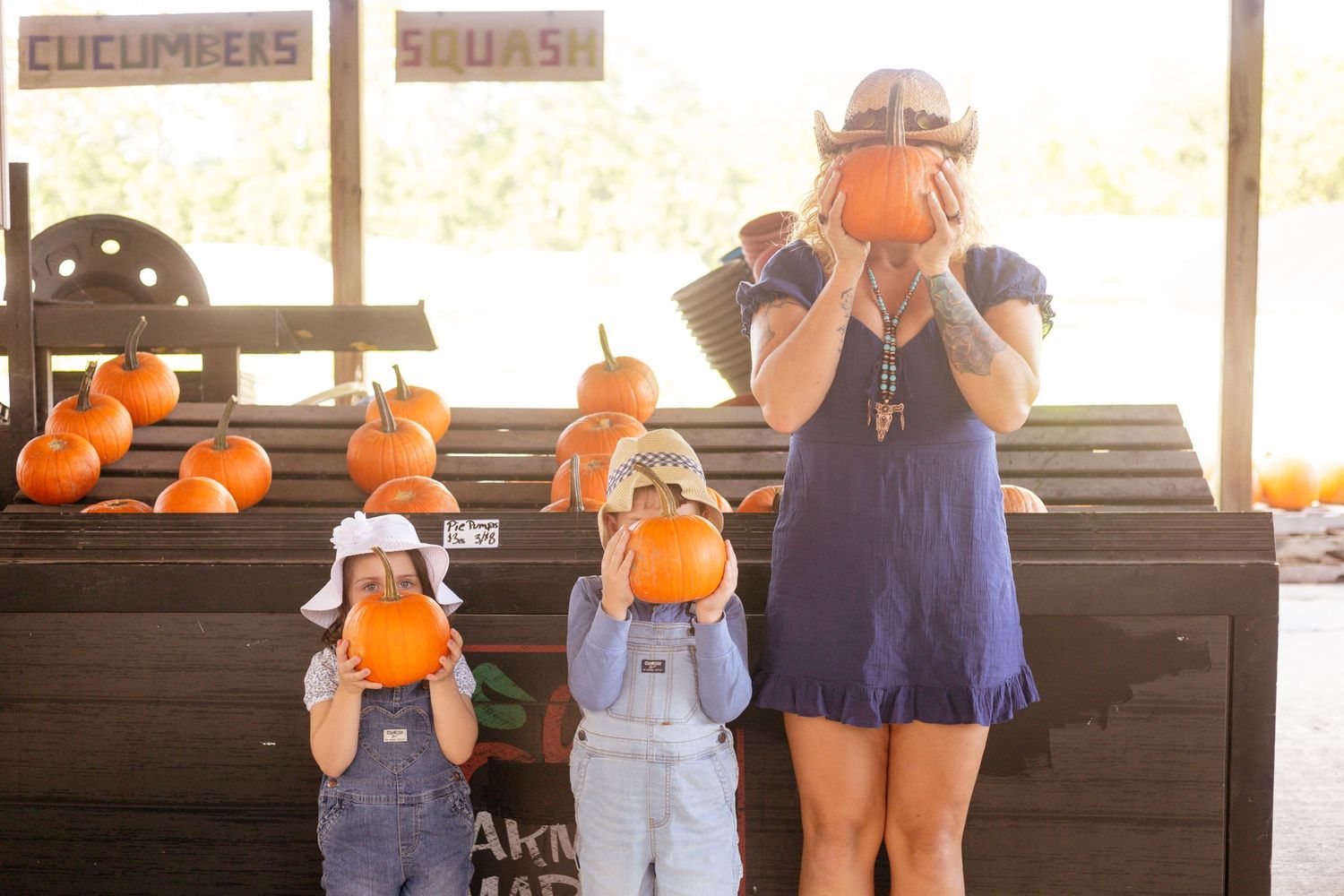 A mother and two children hold pumpkins over their faces at a fall harvest farm stand.
