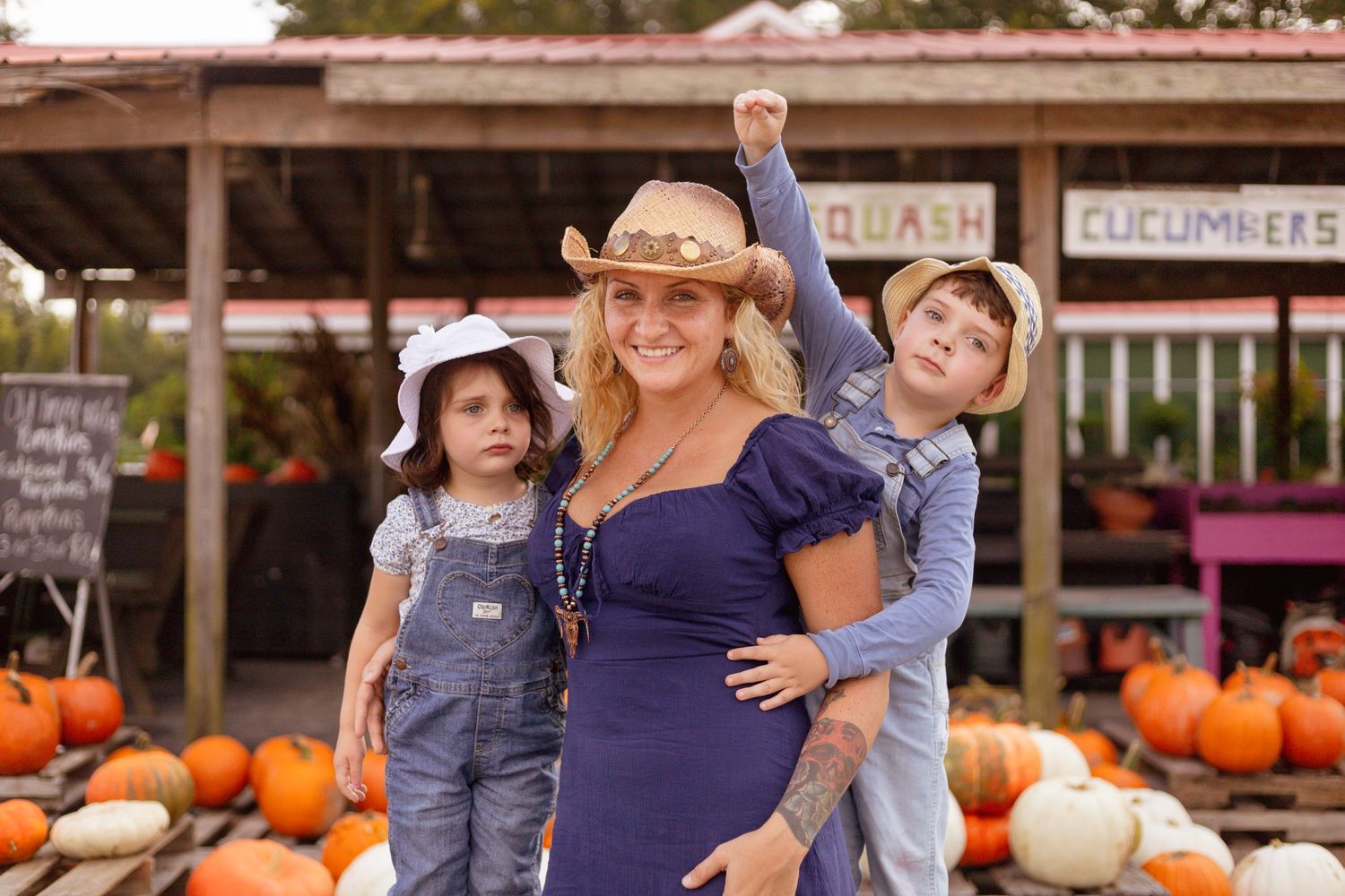 A pregnant woman in a cowboy hat poses with two children at a pumpkin patch.