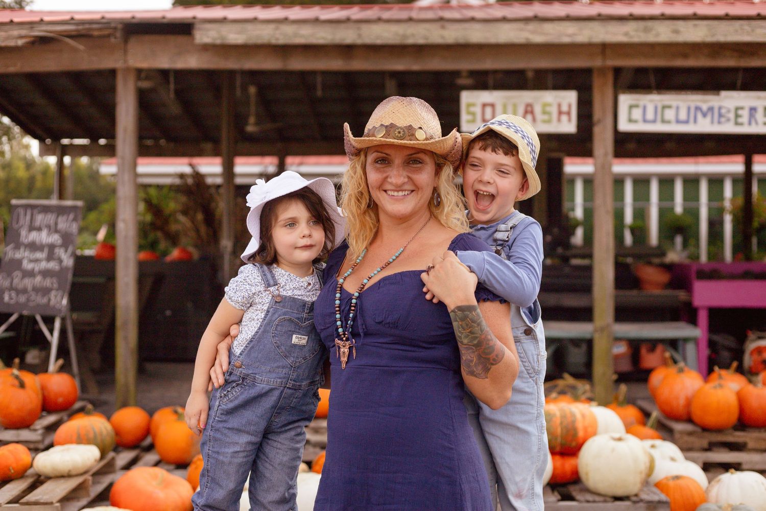 A smiling pregnant woman in a straw hat stands with two children at an outdoor pumpkin patch.