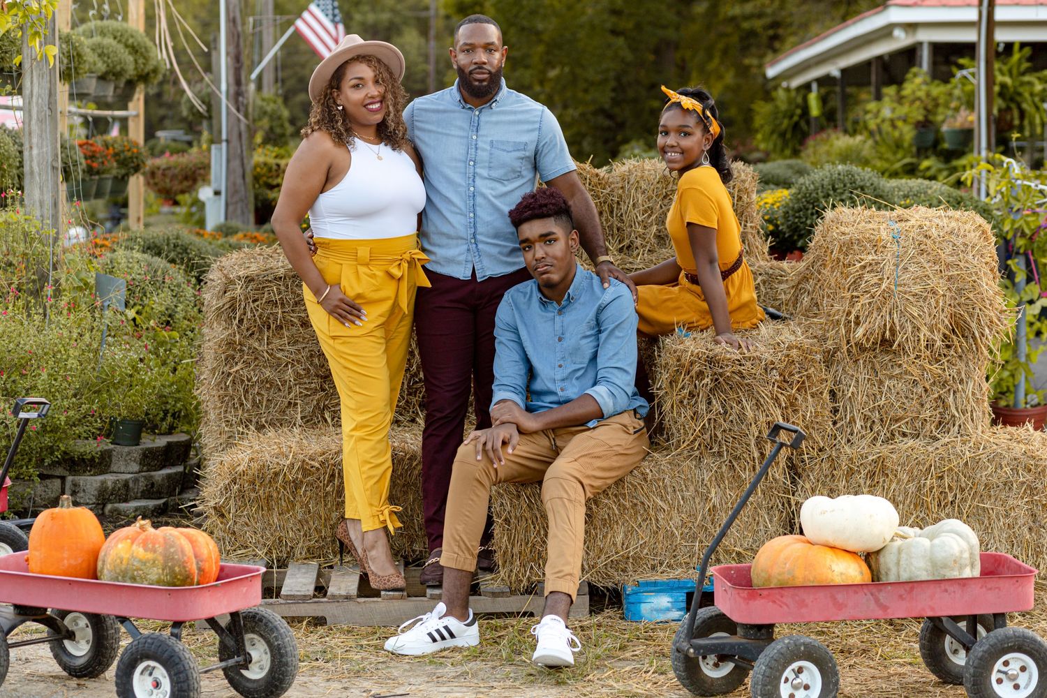 Family of four posing at a fall pumpkin patch with hay bales, pumpkins, and red wagons in the background.