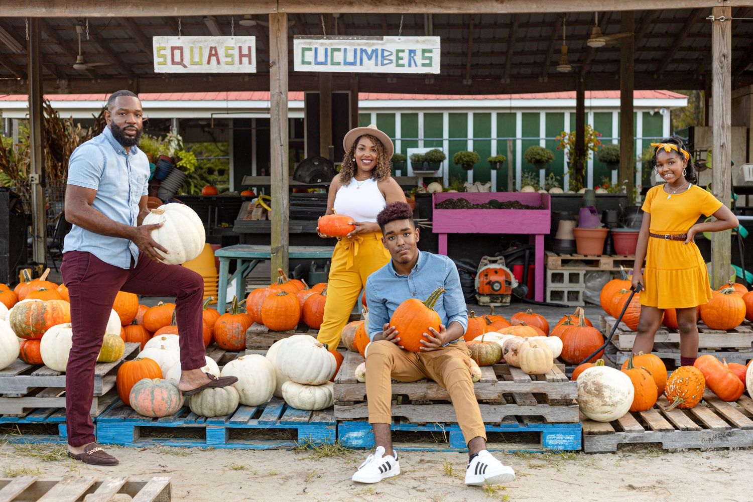 Family of four holding pumpkins at a farm stand with squash and cucumbers signs during a fall photo session.
