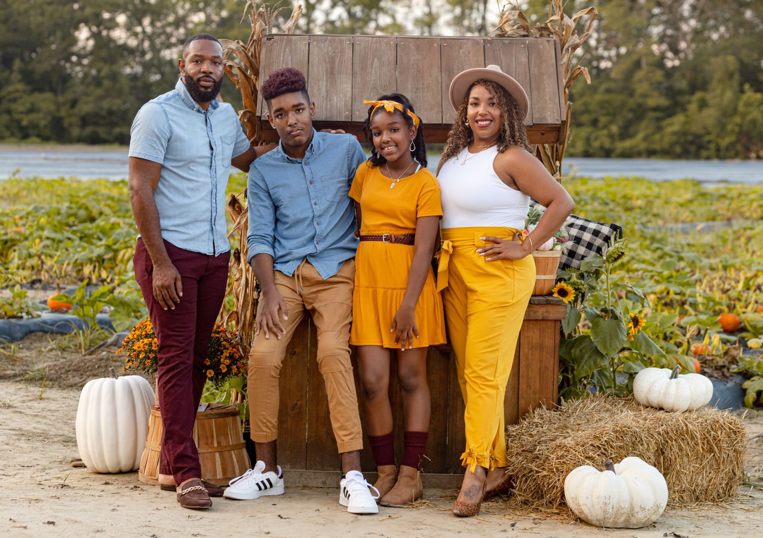 Family of four posing together at a fall pumpkin patch with white pumpkins and rustic wooden decor.