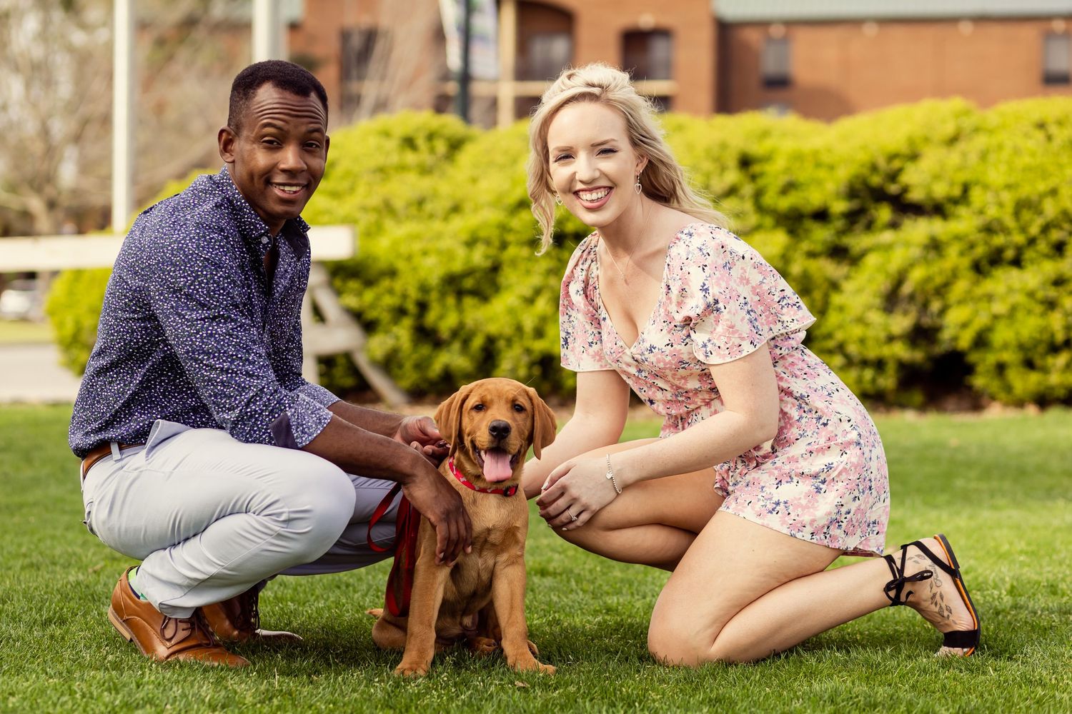 Smiling couple kneeling on grass posing with an adorable golden retriever puppy on a red leash.