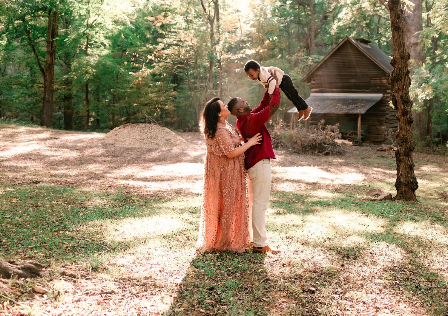 Family of three with parents lifting their child joyfully in a sunlit wooded outdoor setting.