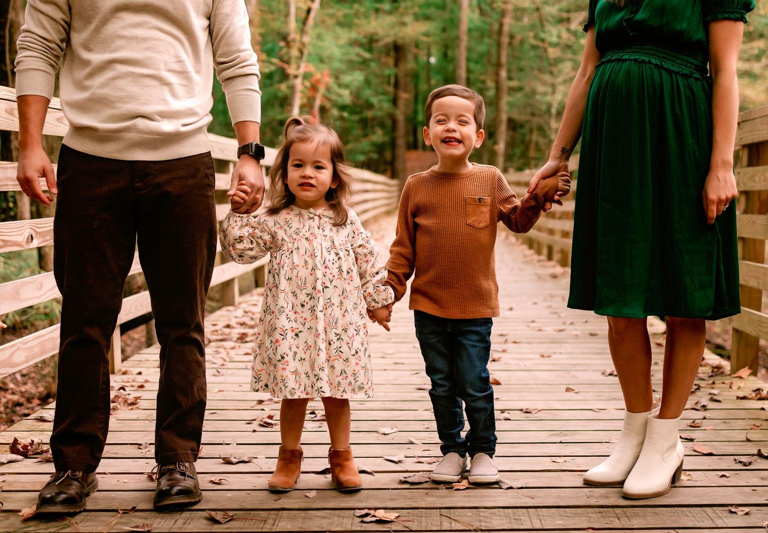 Two young children holding parents hands while walking on a wooden bridge in an autumn forest.