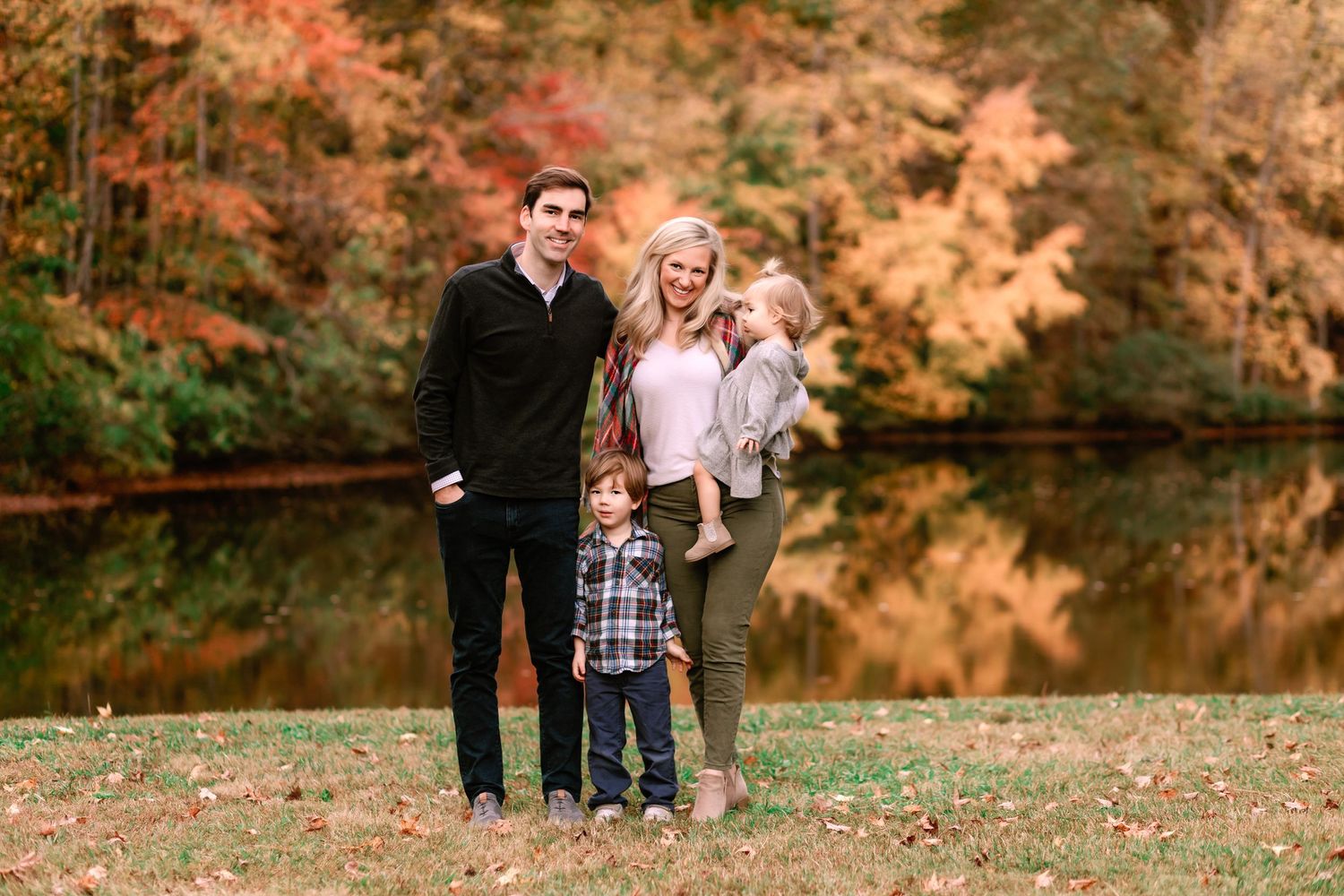 Family of four posing by a reflective pond surrounded by vibrant orange and red fall foliage.