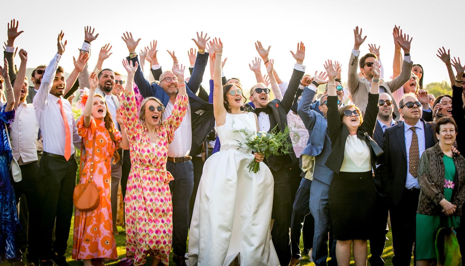 Large group of wedding guests raise their hands in celebration outdoors.