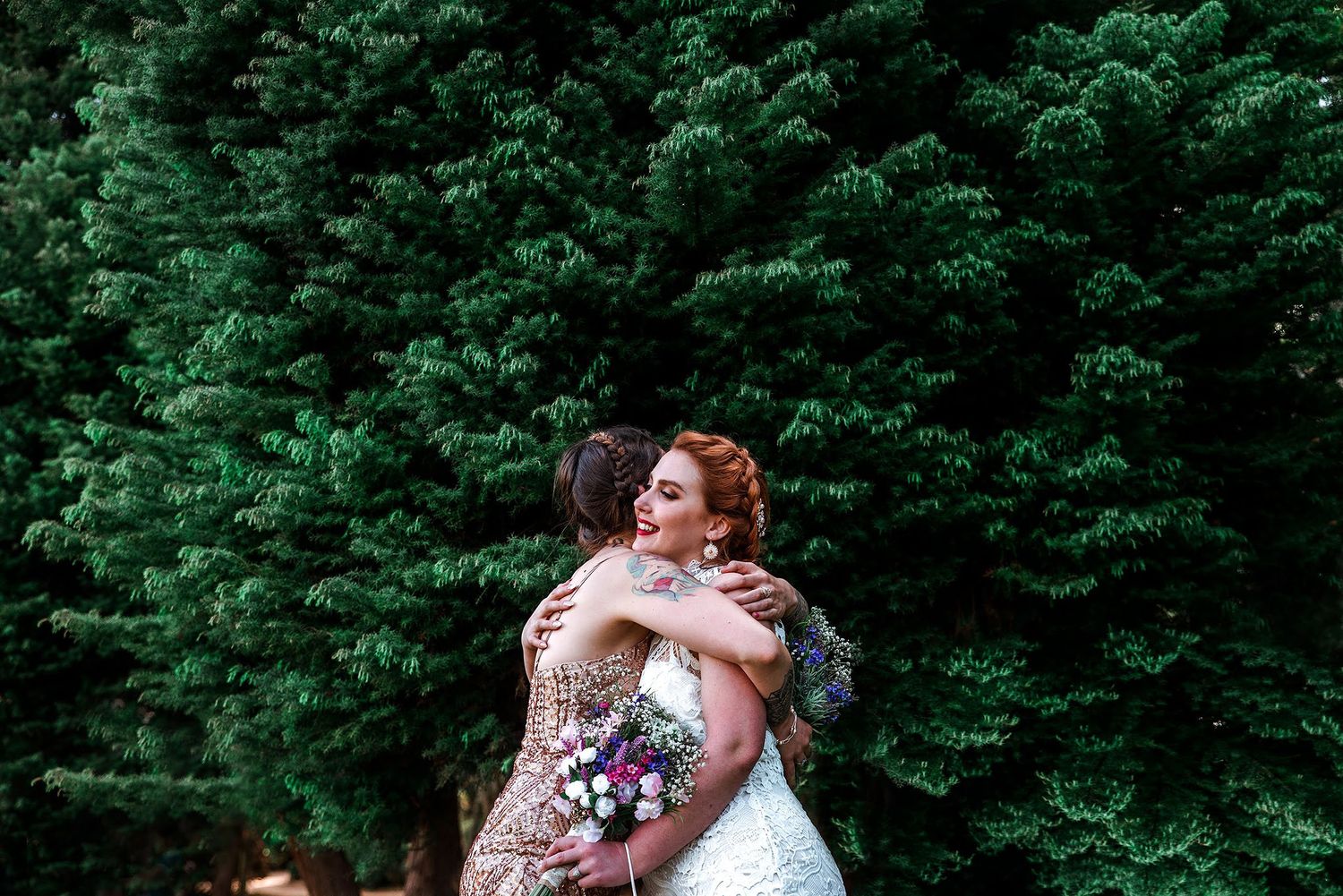 Two people in wedding attire embrace in front of a green garden backdrop.