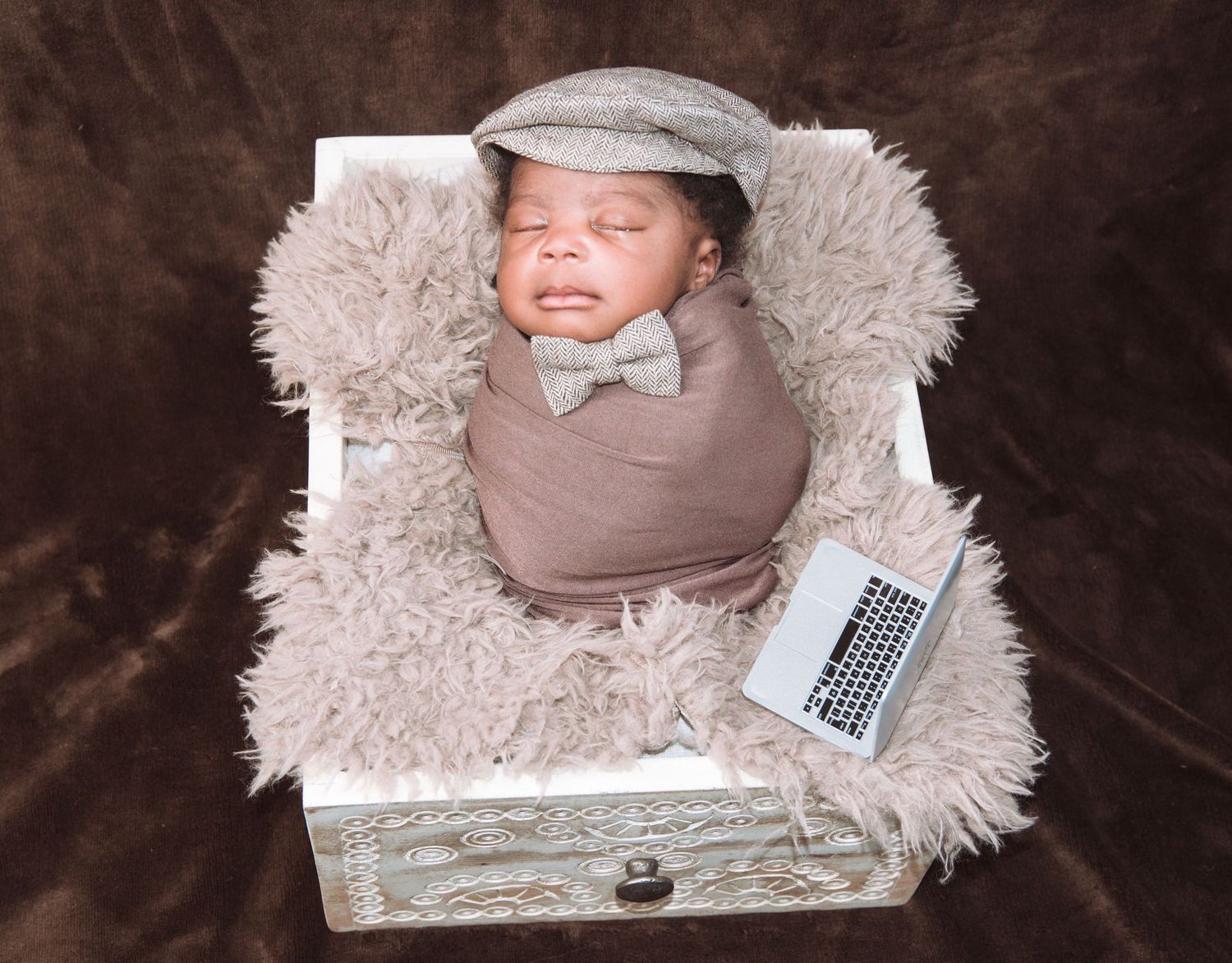 A baby wrapped in grey with a newsboy cap poses with a laptop on fuzzy blankets.