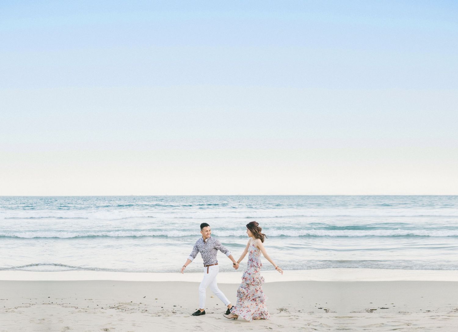 A couple runs joyfully along a serene beach with white sand and ocean waves under a bright sky.
