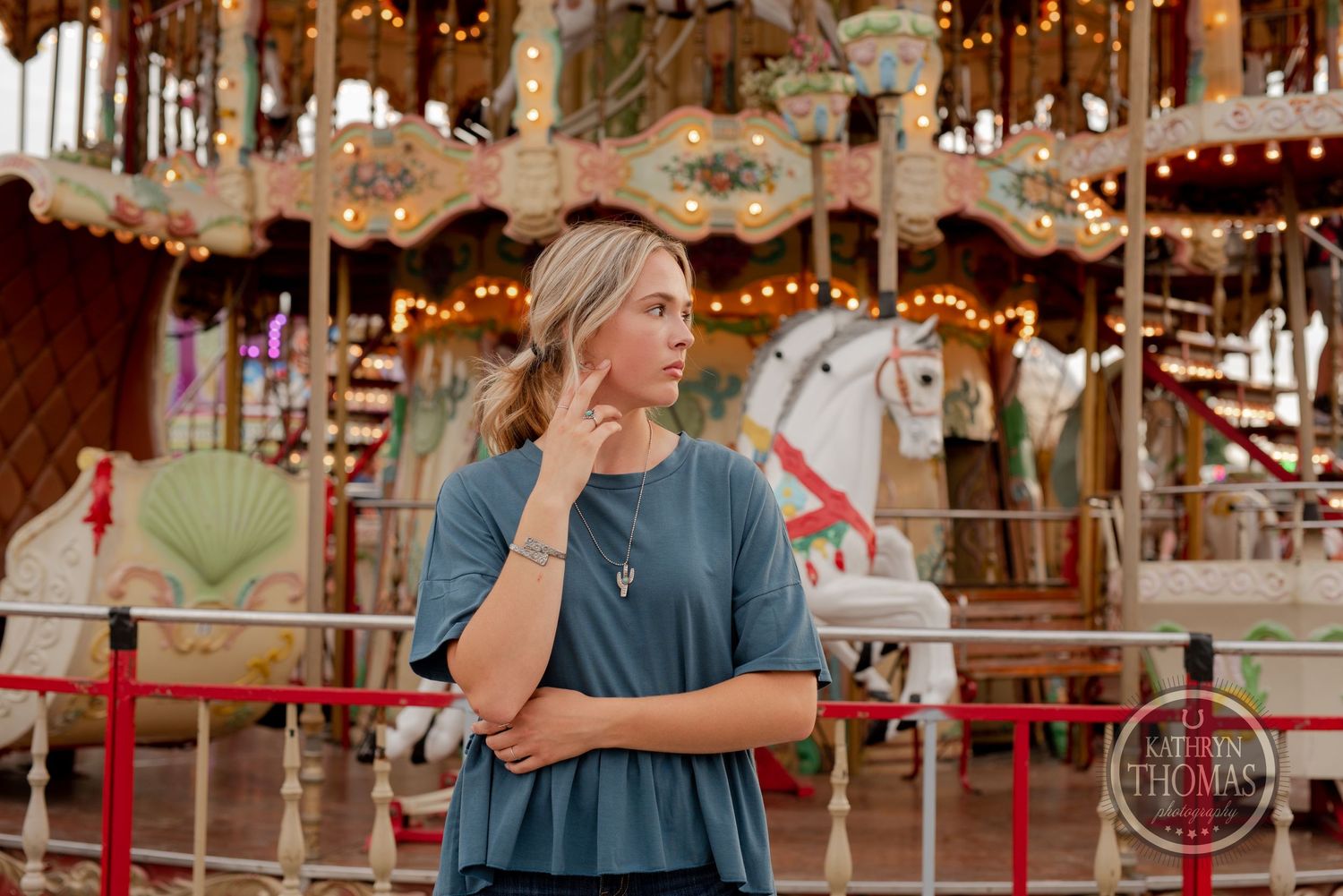 Person in casual blue top standing before vintage carousel with glowing lights.