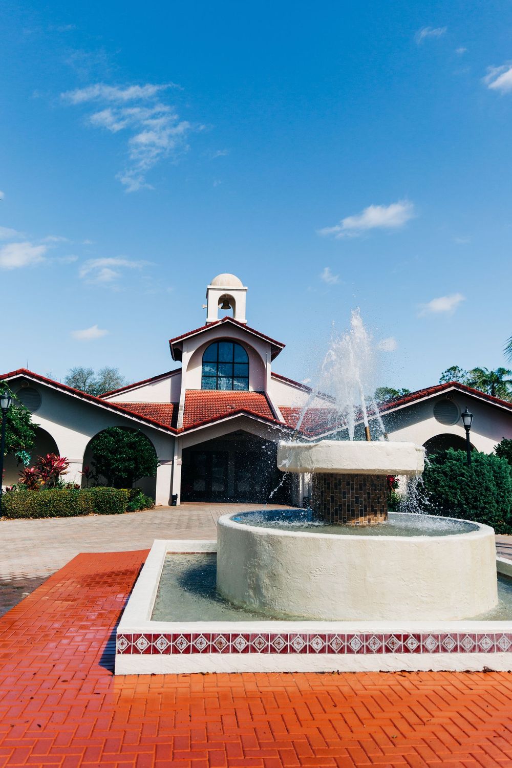 Spanish style church exterior with red brick courtyard and central water fountain under bright blue sky.
